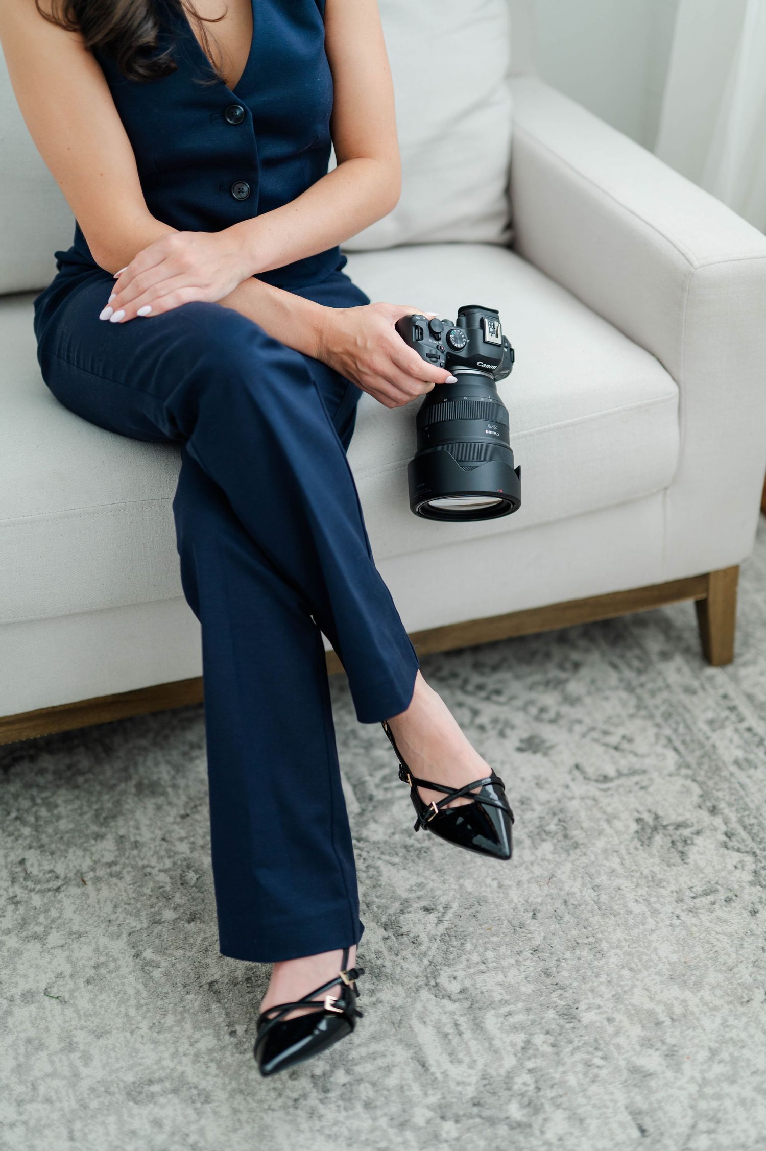 Close-up view of navy jumpsuit and black patent leather heels on white couch with gray patterned rug below.