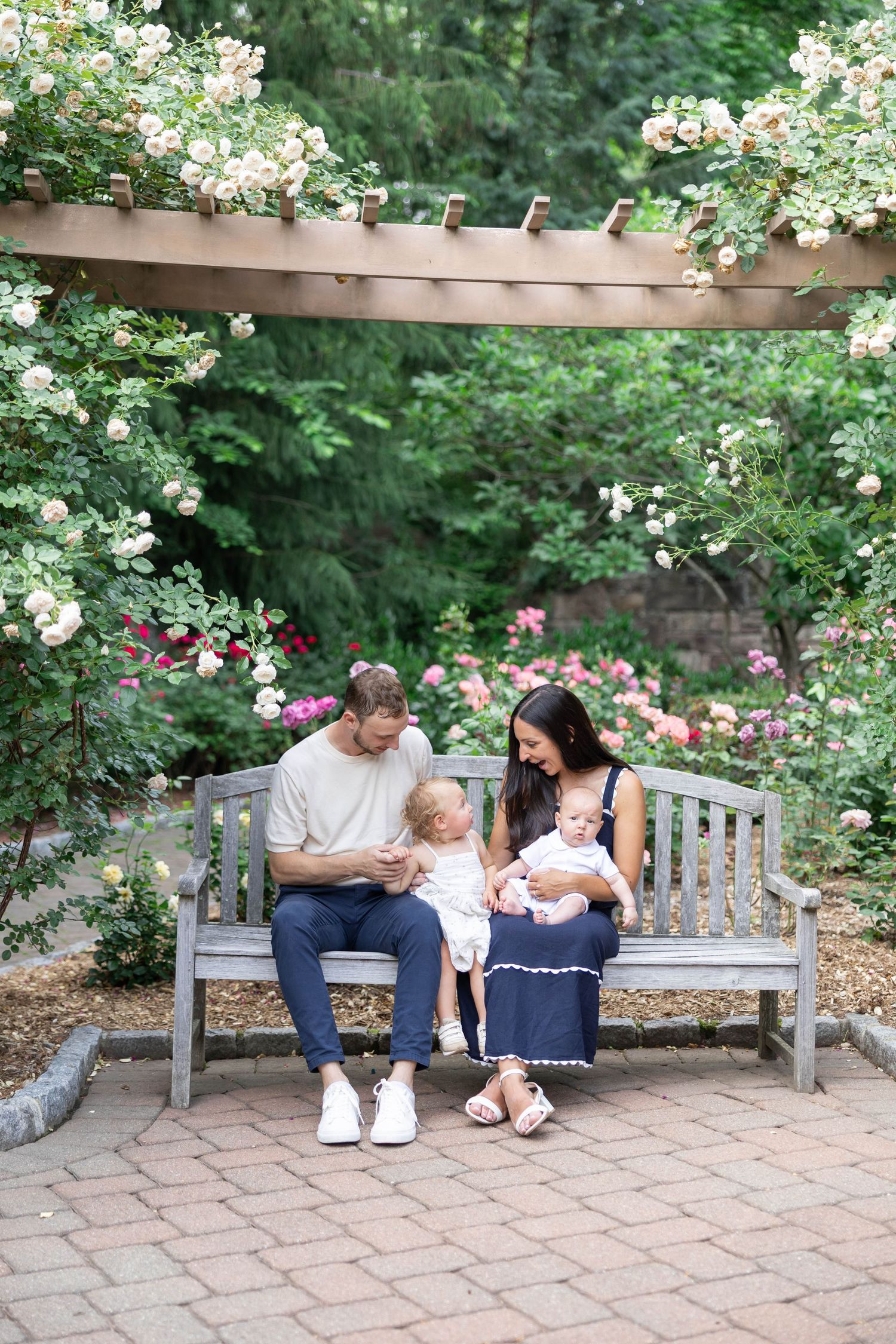 Family sitting together on wooden bench in garden surrounded by blooming white and pink roses.