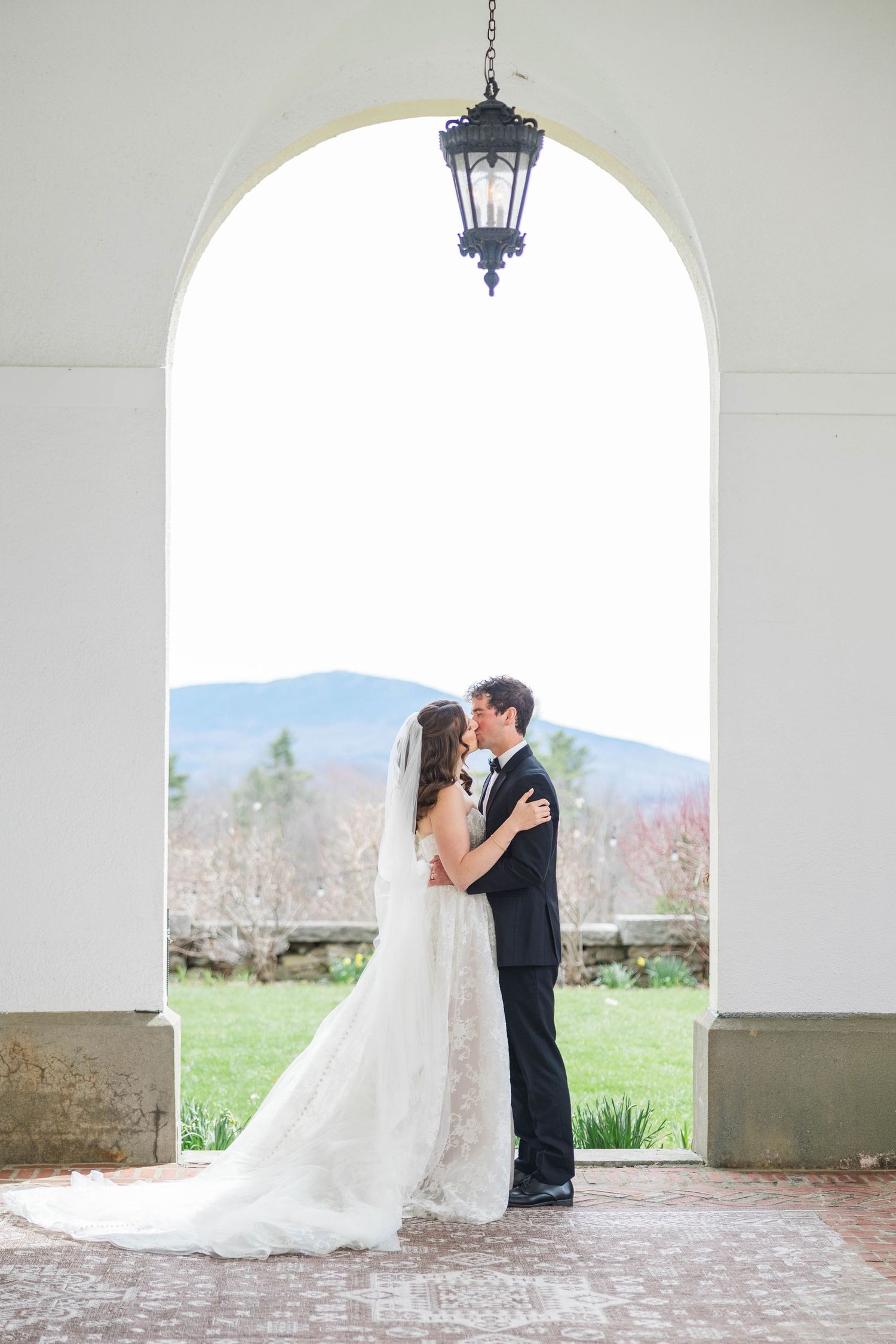 Bride and groom sharing a kiss under an elegant white arch with a hanging lantern, mountains visible in the background.
