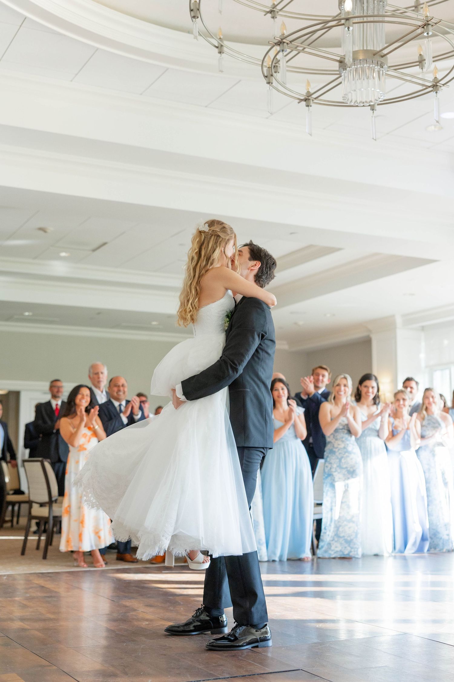 Groom lifts bride during first dance at elegant wedding reception with bridesmaids in light blue gowns applauding.