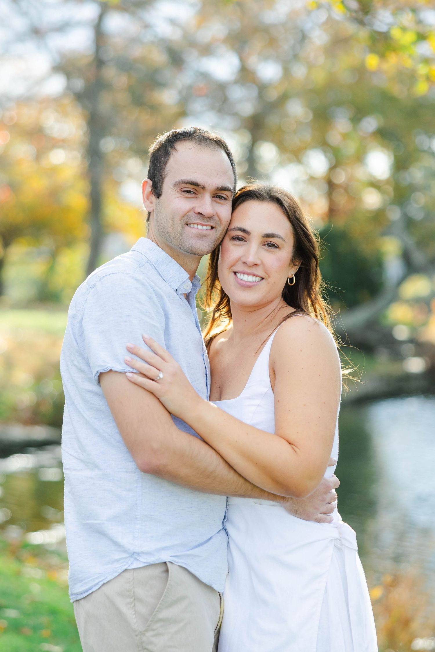 A couple shares a joyful embrace outdoors on a sunny autumn day with blurred trees and water in the background.
