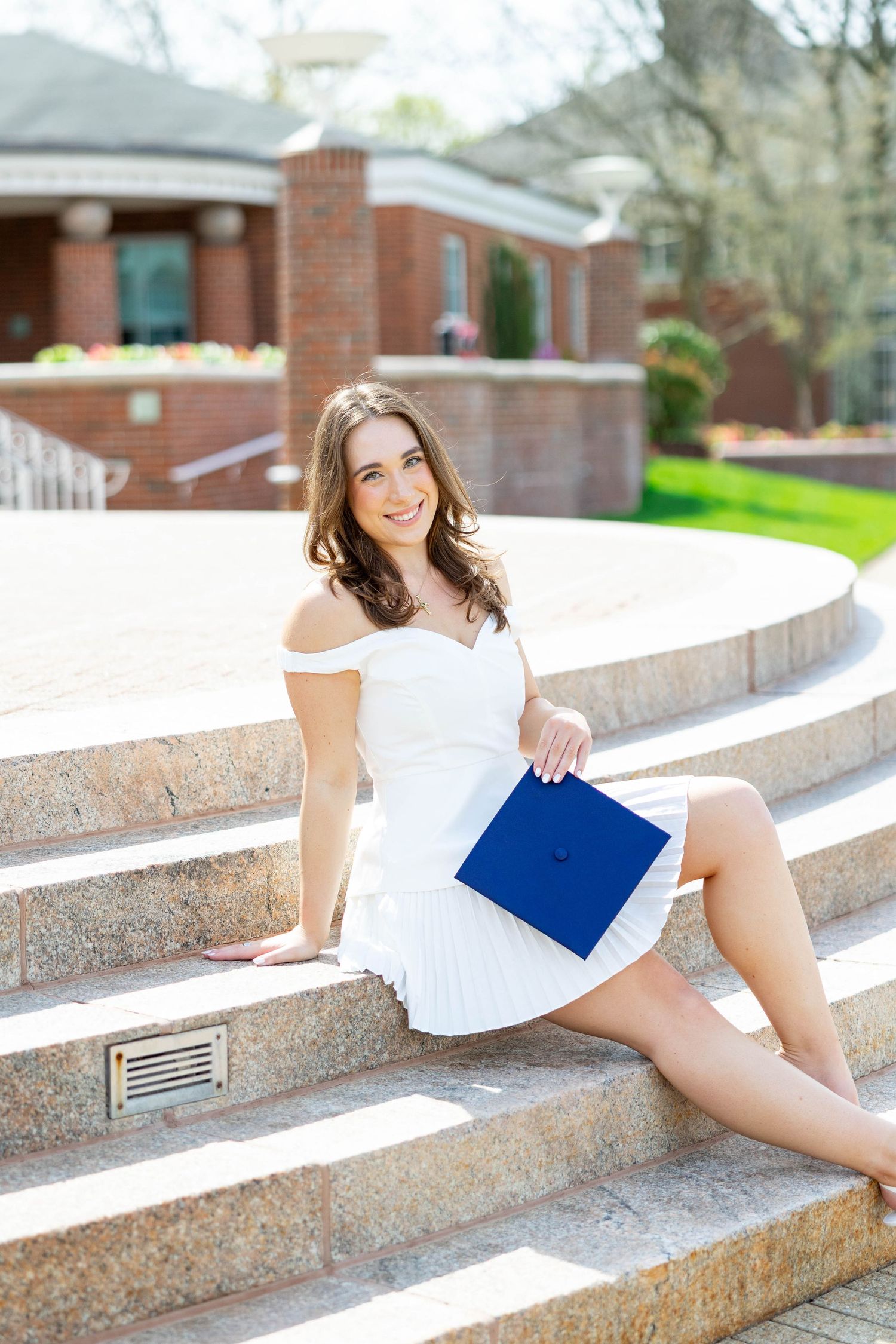 Young woman in white dress holding blue graduation cap, sitting on stone steps outside brick building.