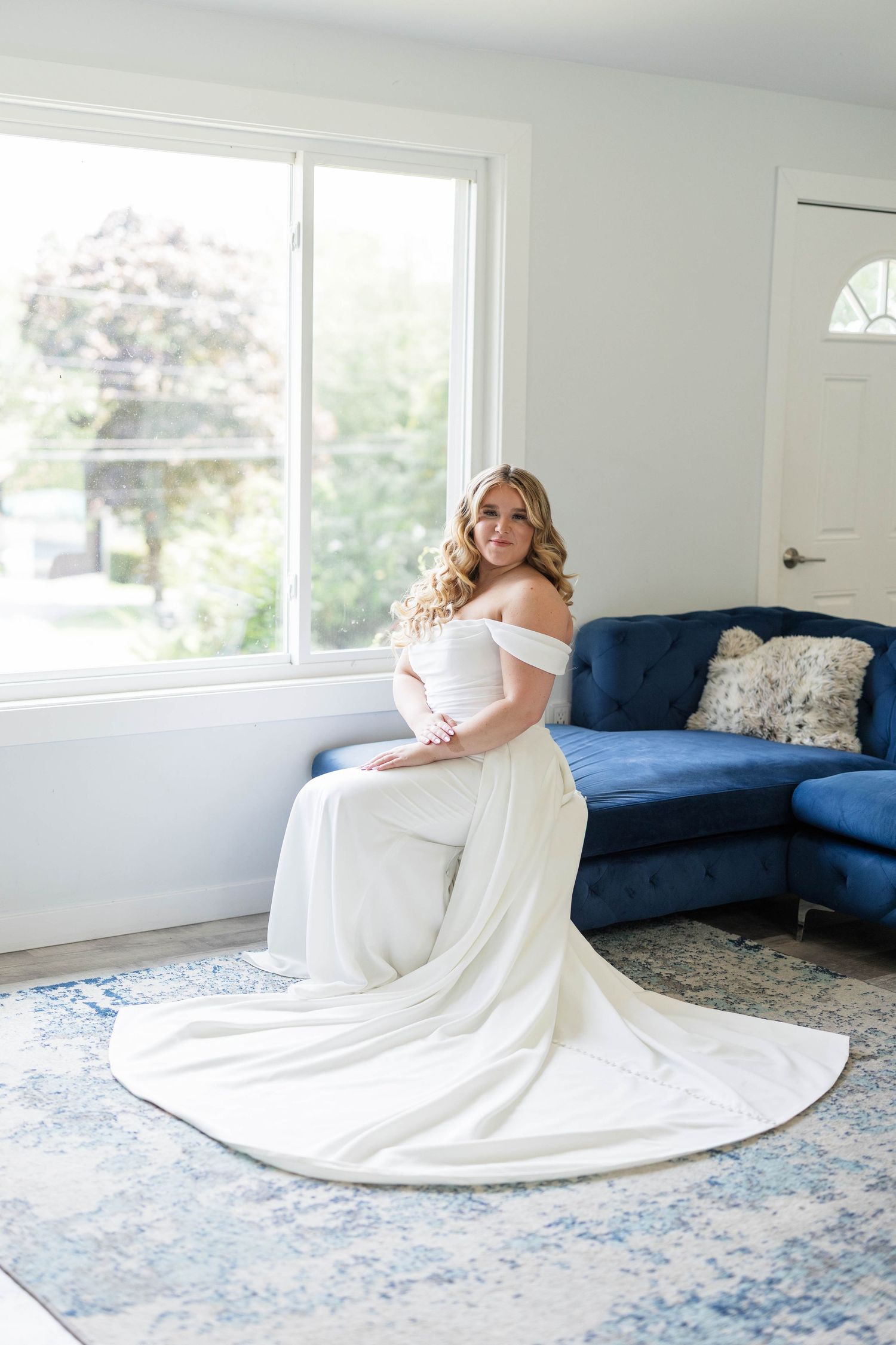 A bride in an elegant off-shoulder white gown with a long train sits gracefully near a blue sofa in a bright room.