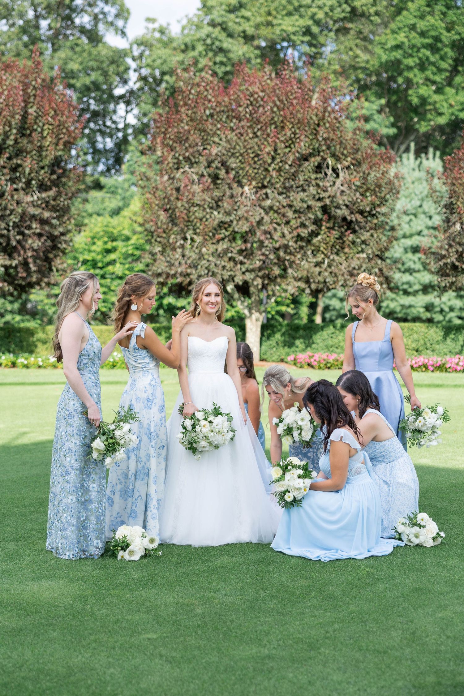 A group in light blue dresses and white gown gather on a lush green lawn for wedding party photos.