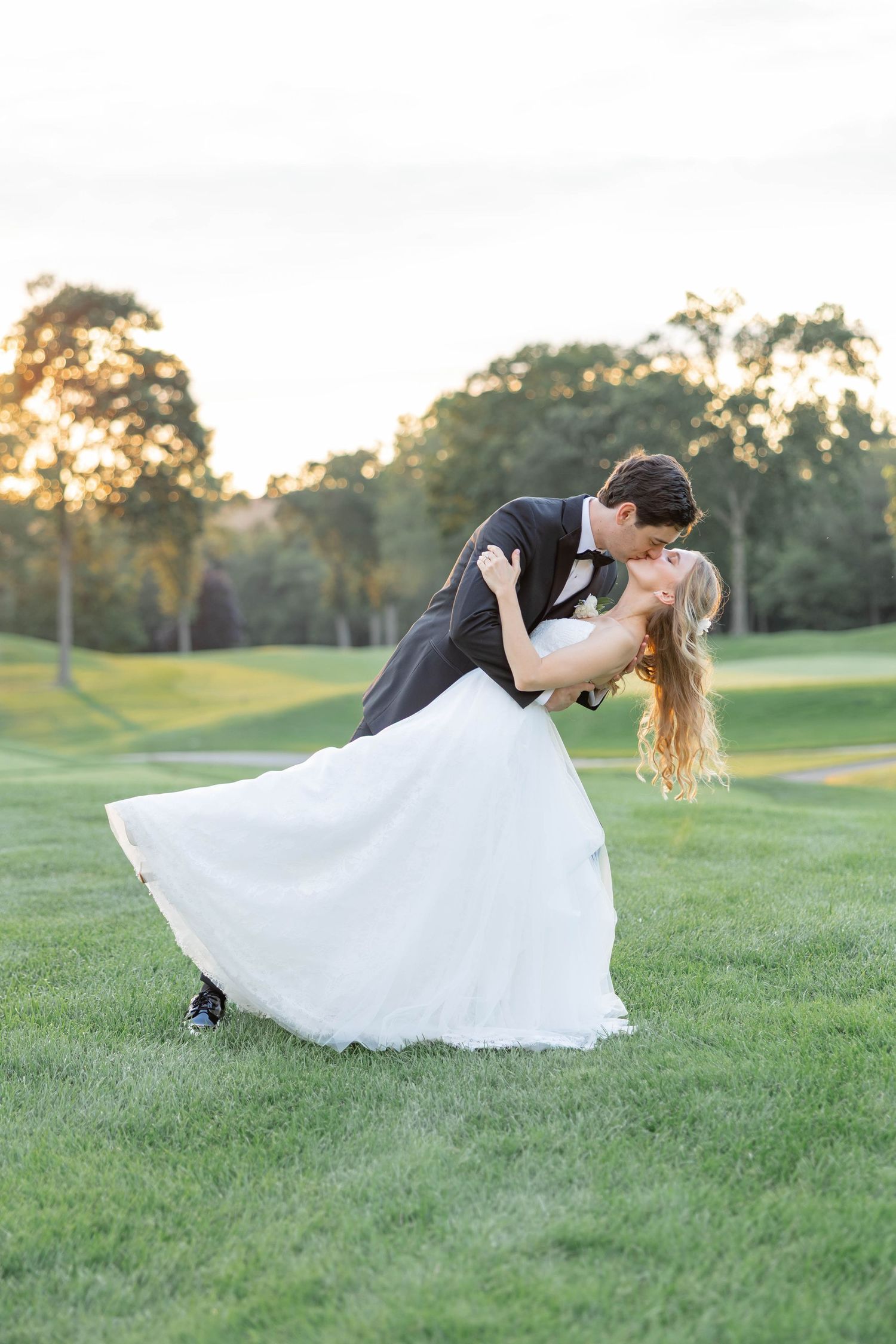 Bride and groom share a romantic dip kiss on lush green golf course at golden hour sunset.