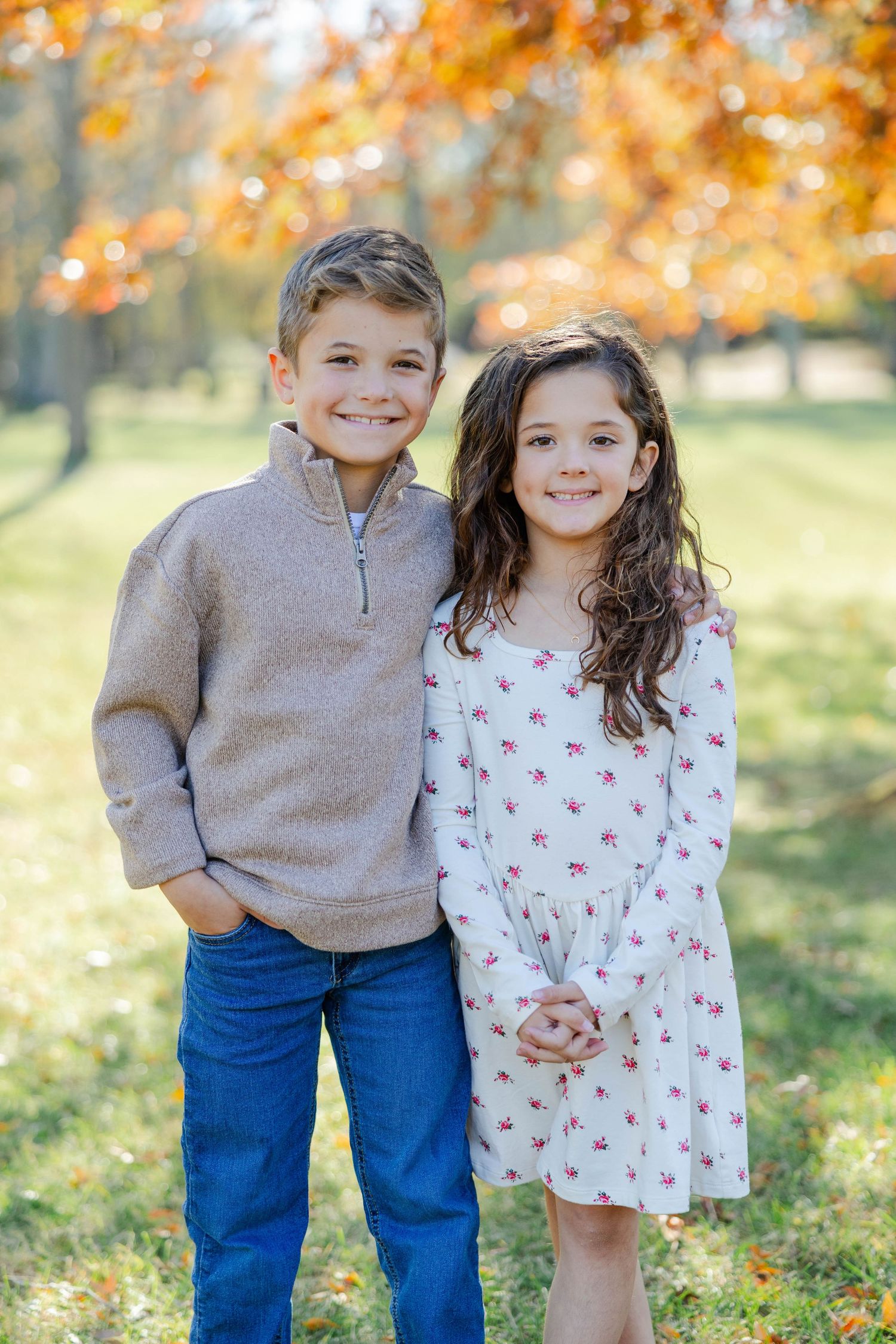 Two children stand together outdoors in autumn with golden leaves and bokeh lighting in the background.
