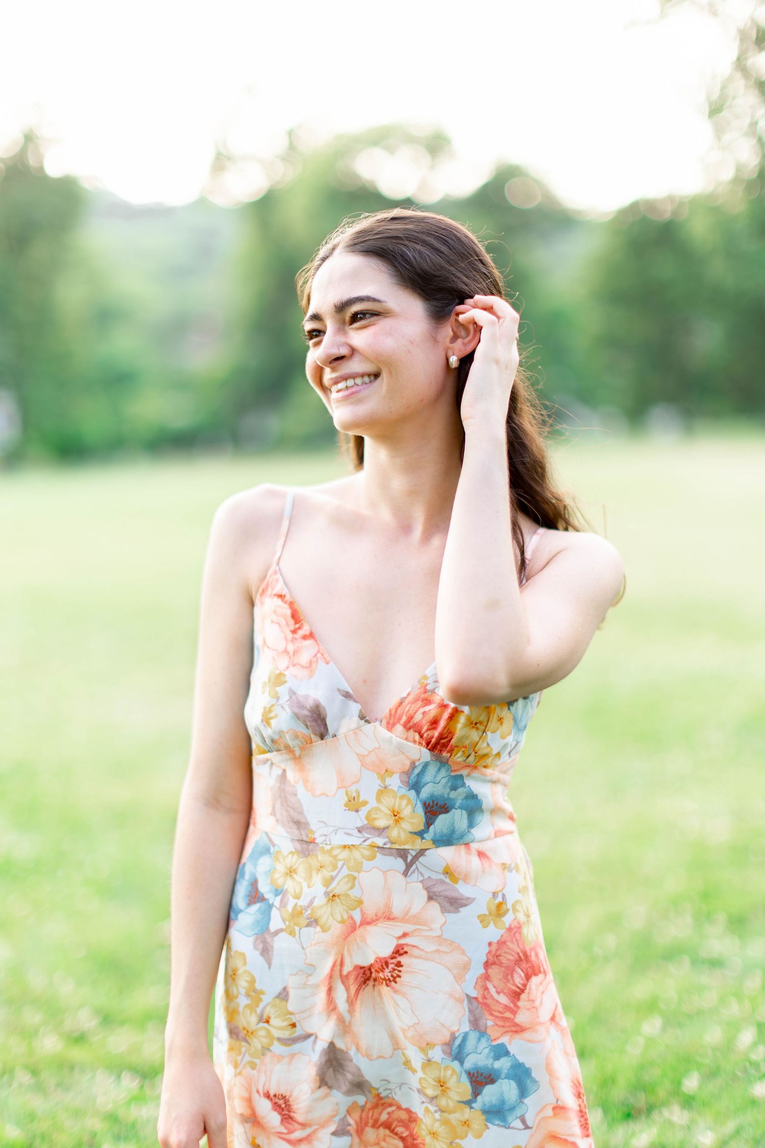 Smiling young woman in a floral sundress standing in a green field, tucking hair behind her ear on a sunny day.