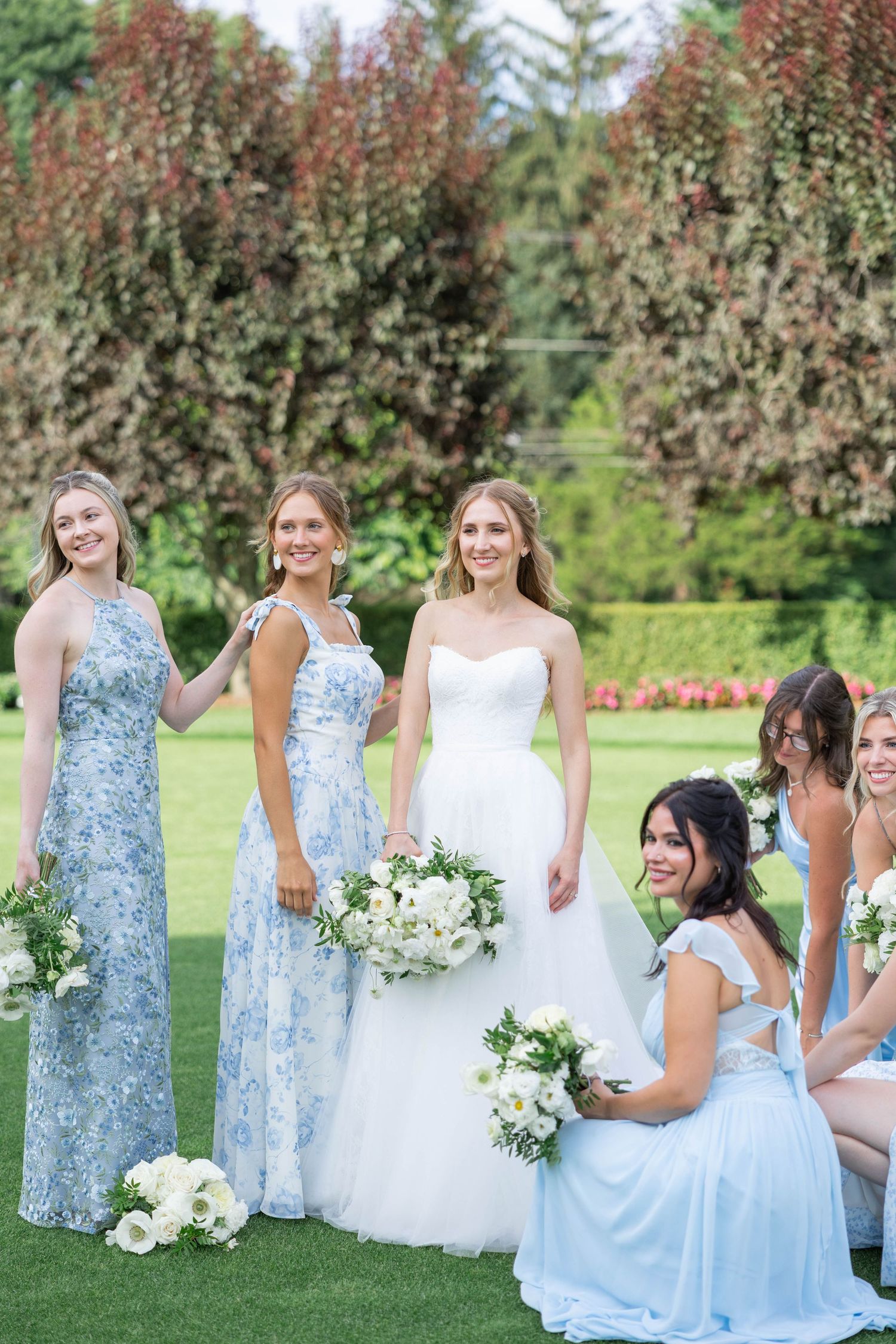 A bridal party in light blue bridesmaid dresses and white wedding gown poses together on a garden lawn.