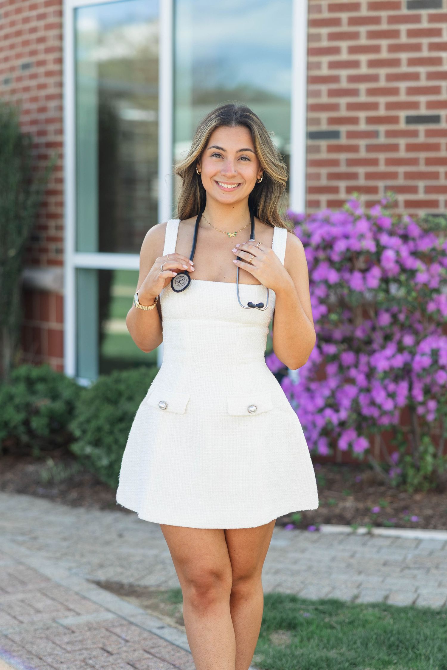 Smiling young woman in white mini dress holding sunglasses, posing outdoors near brick building with purple flowers.