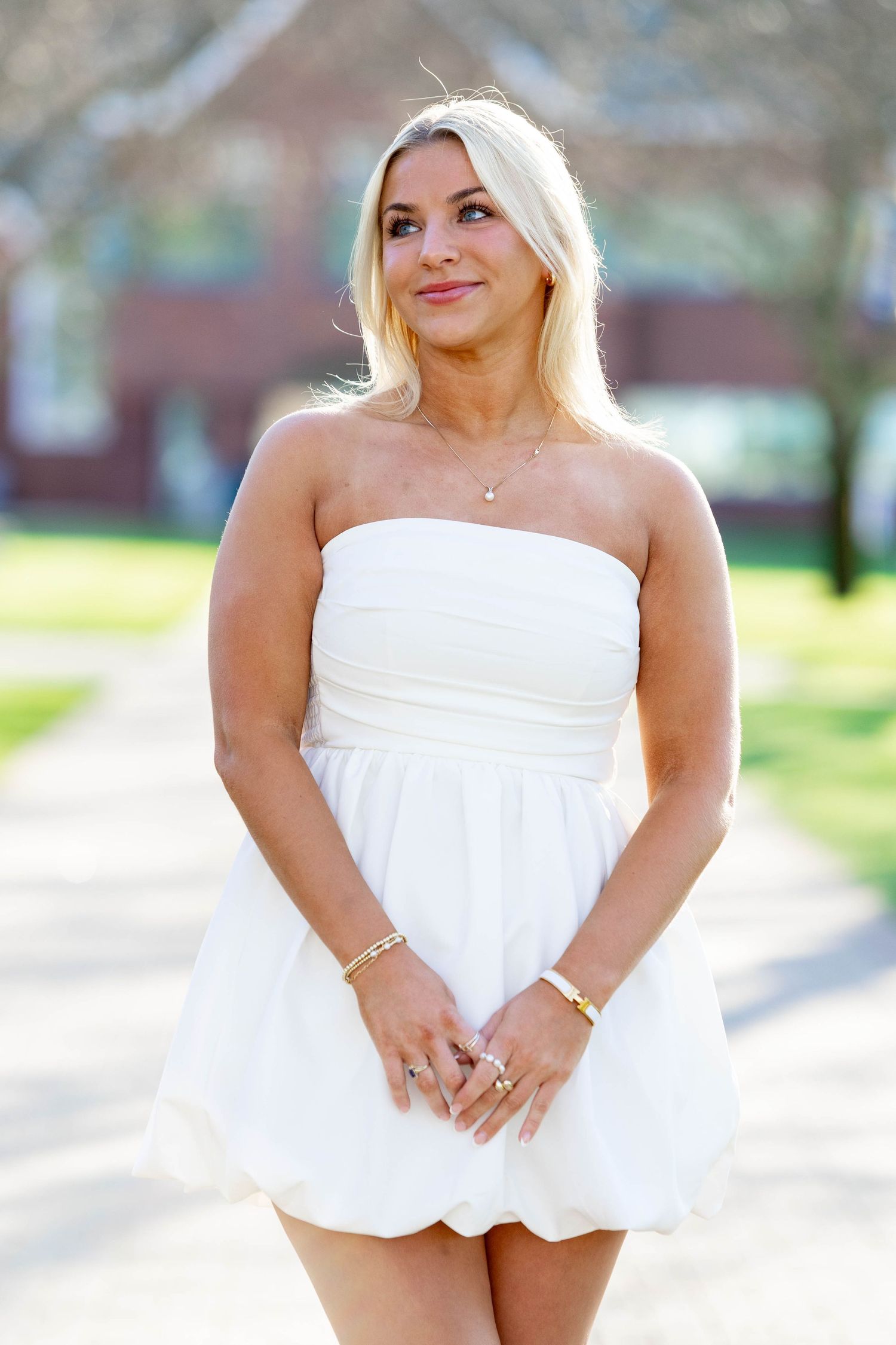 Blonde woman in white strapless mini dress with gold jewelry smiling outdoors on sunny day.