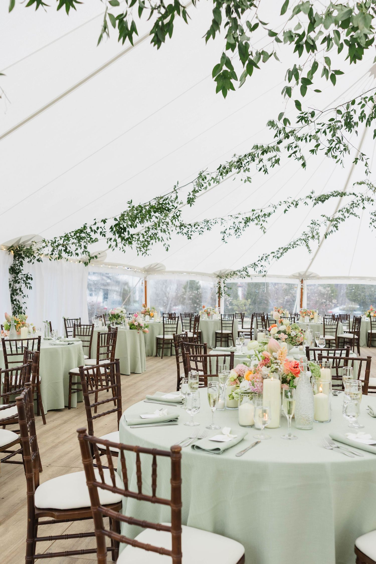 Elegant wedding reception tent with sage green tablecloths, chiavari chairs, floral centerpieces, and lush greenery draped overhead.
