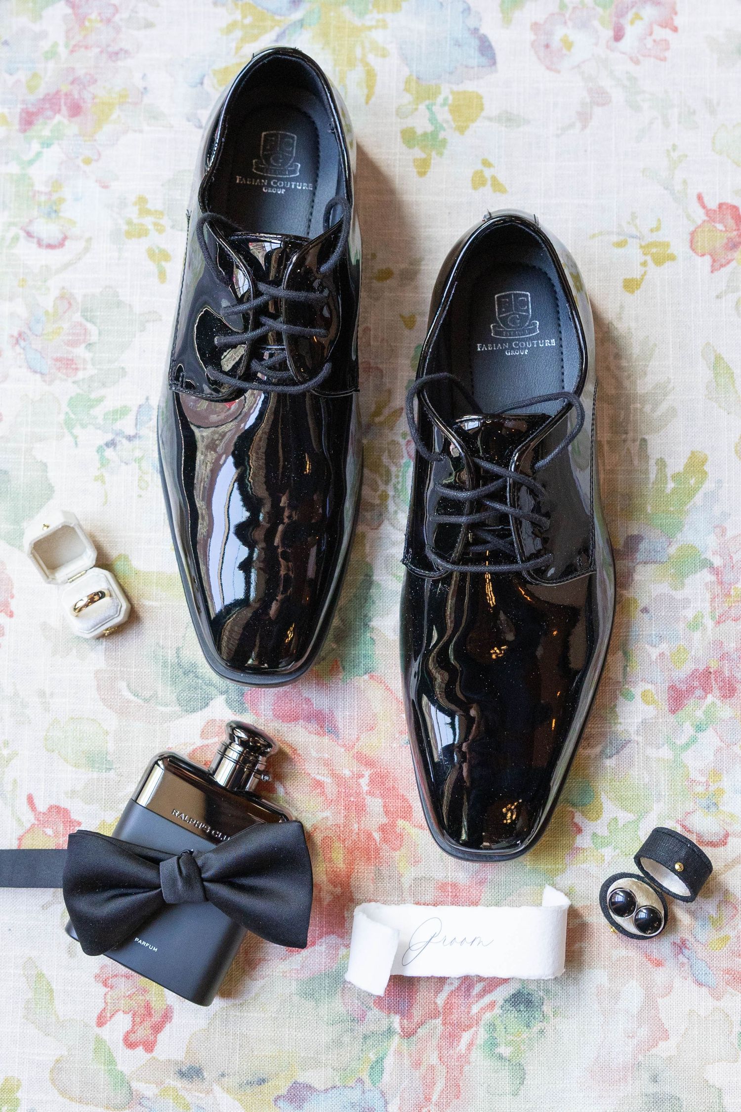 Elegant black patent leather dress shoes with a bow tie and rings arranged on a floral background.