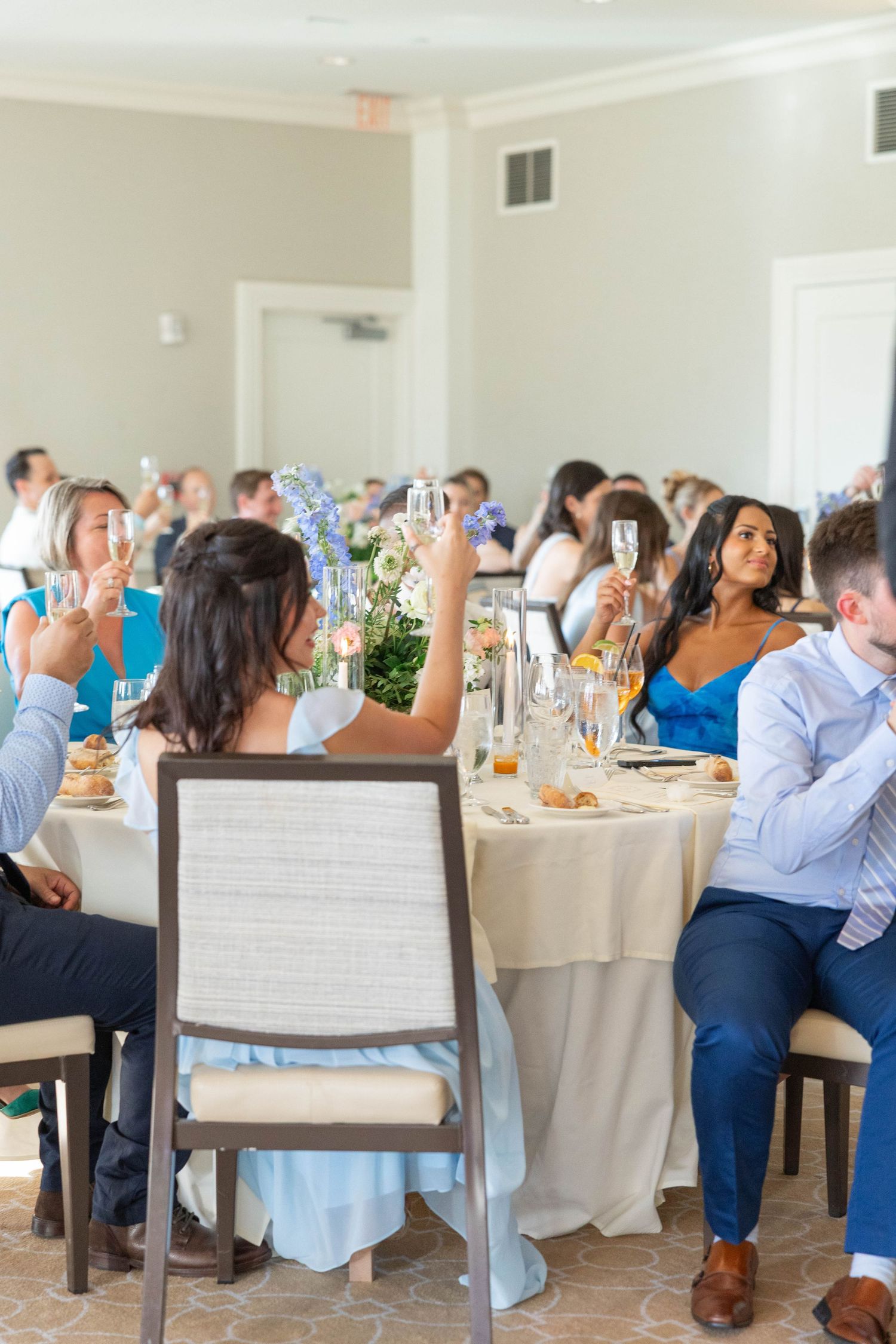 Wedding reception guests seated at round tables with white linens raise their glasses for a celebratory toast.