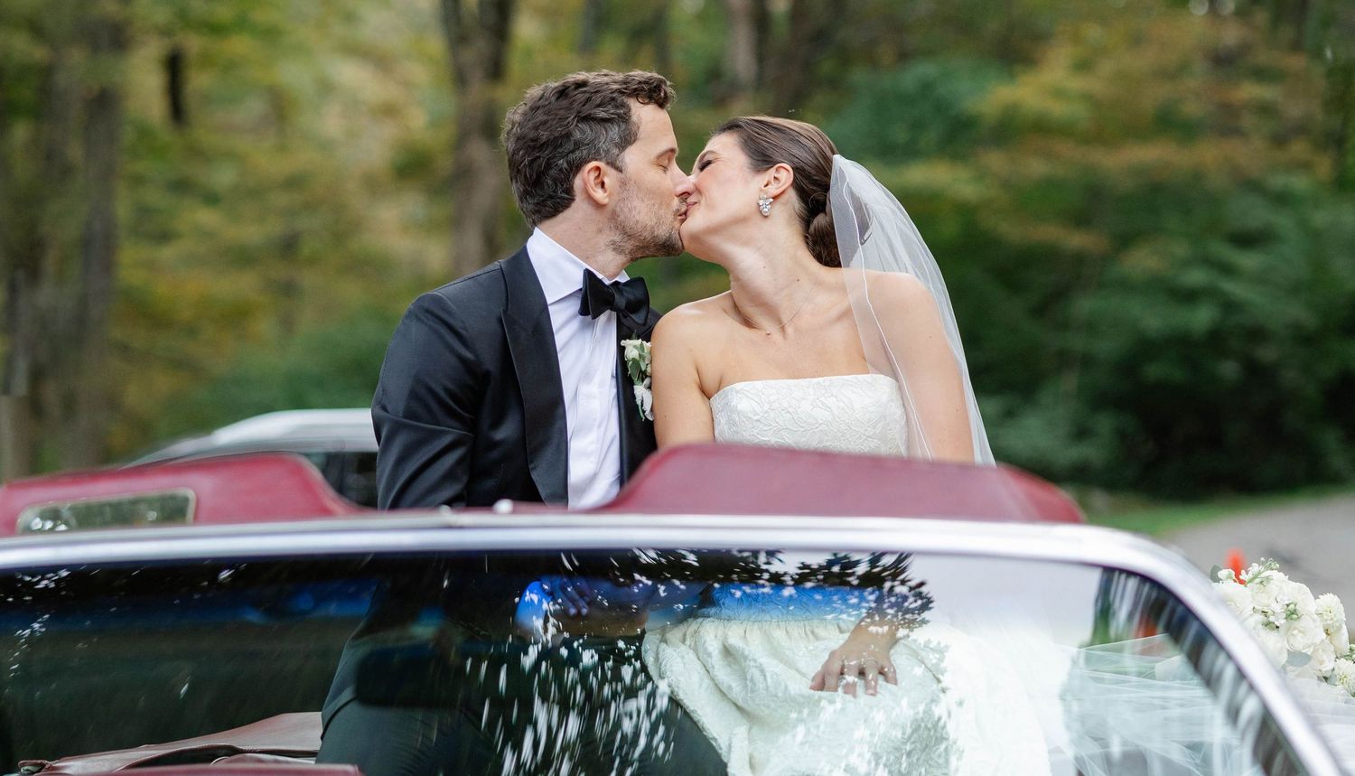 A romantic wedding couple shares a kiss while sitting in a vintage red car decorated with flowers against a forest backdrop.