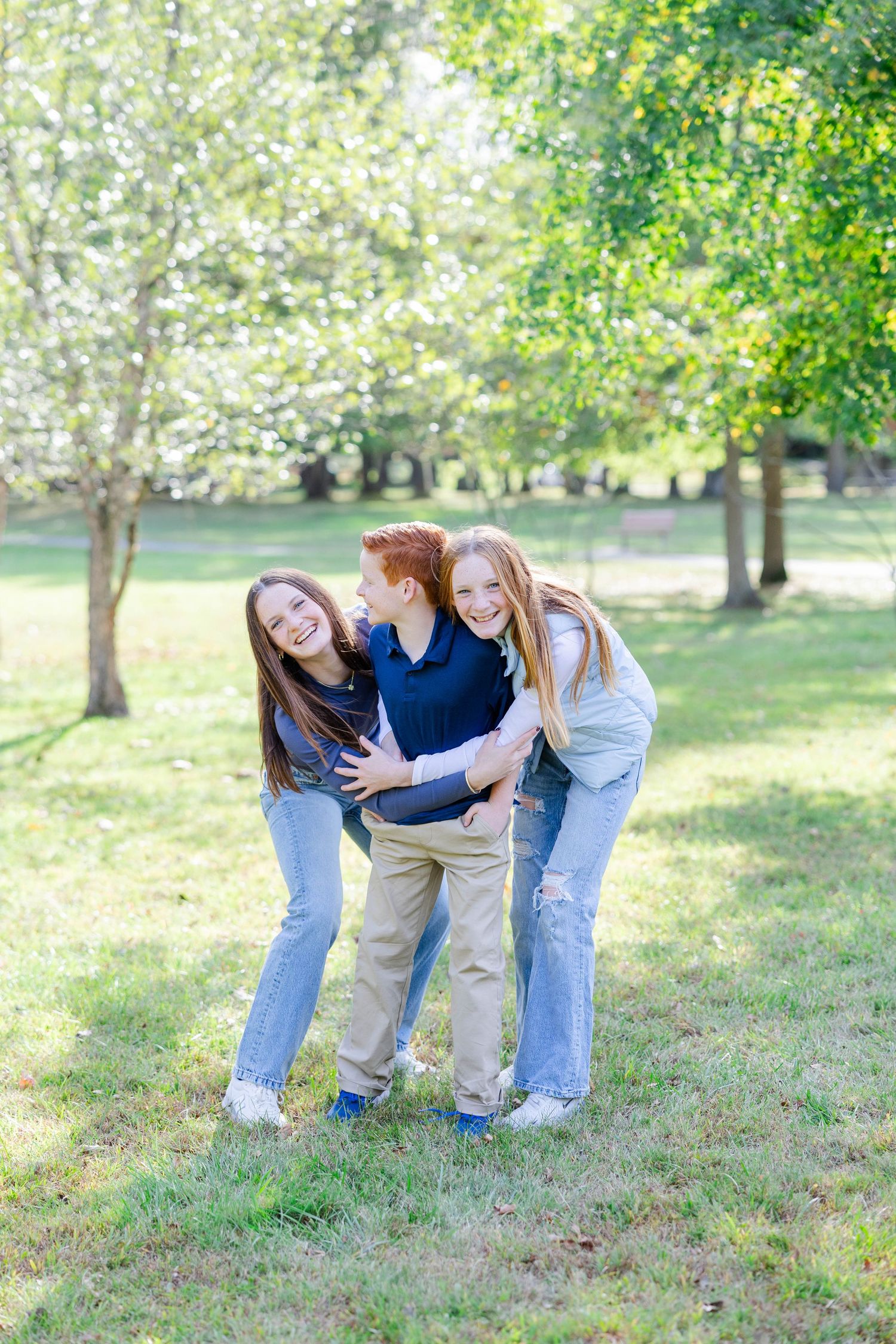 Three people playing and laughing together outdoors on a sunny day in a grassy park setting.