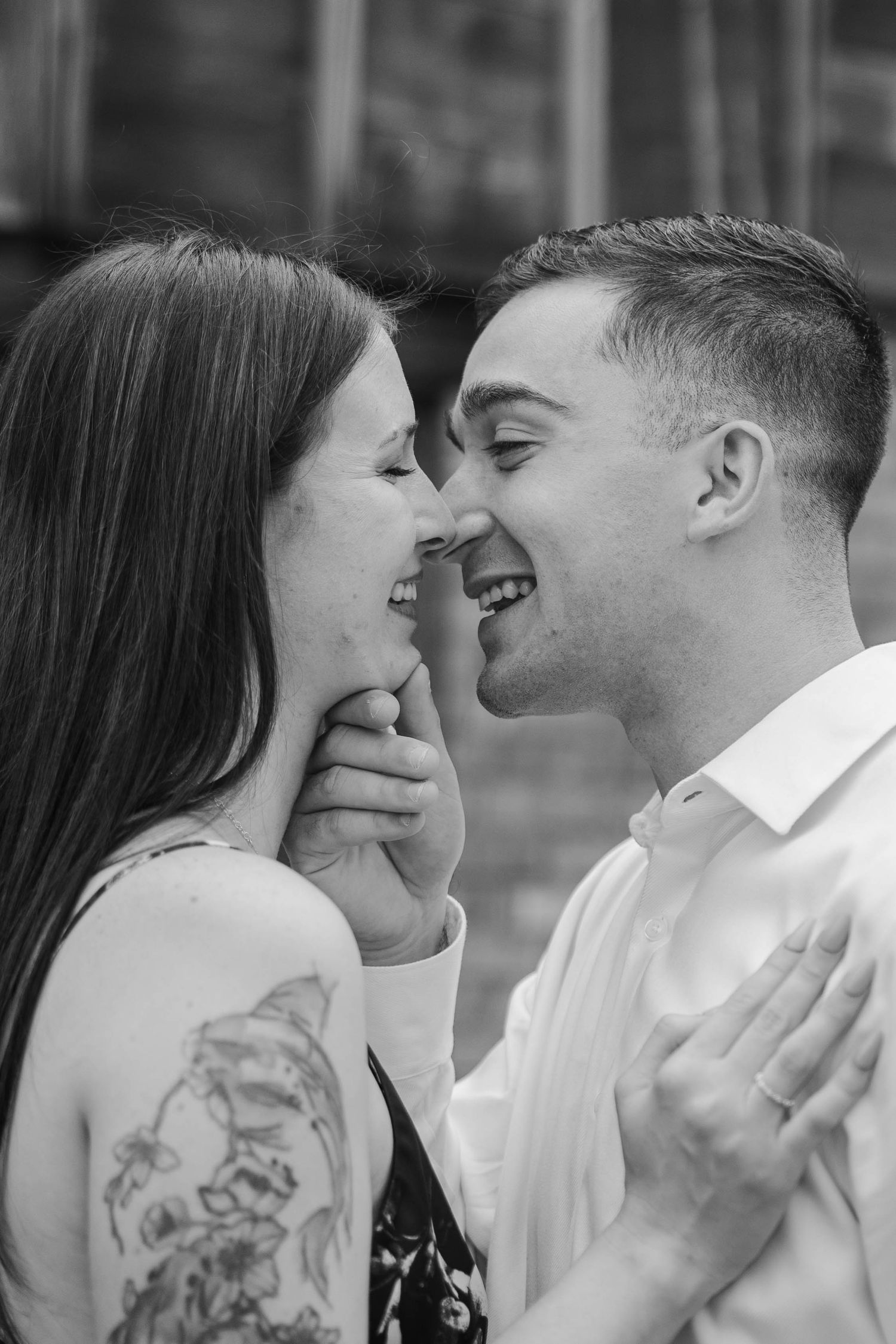 Black and white photo of a smiling couple holding each other close, woman with floral arm tattoo.