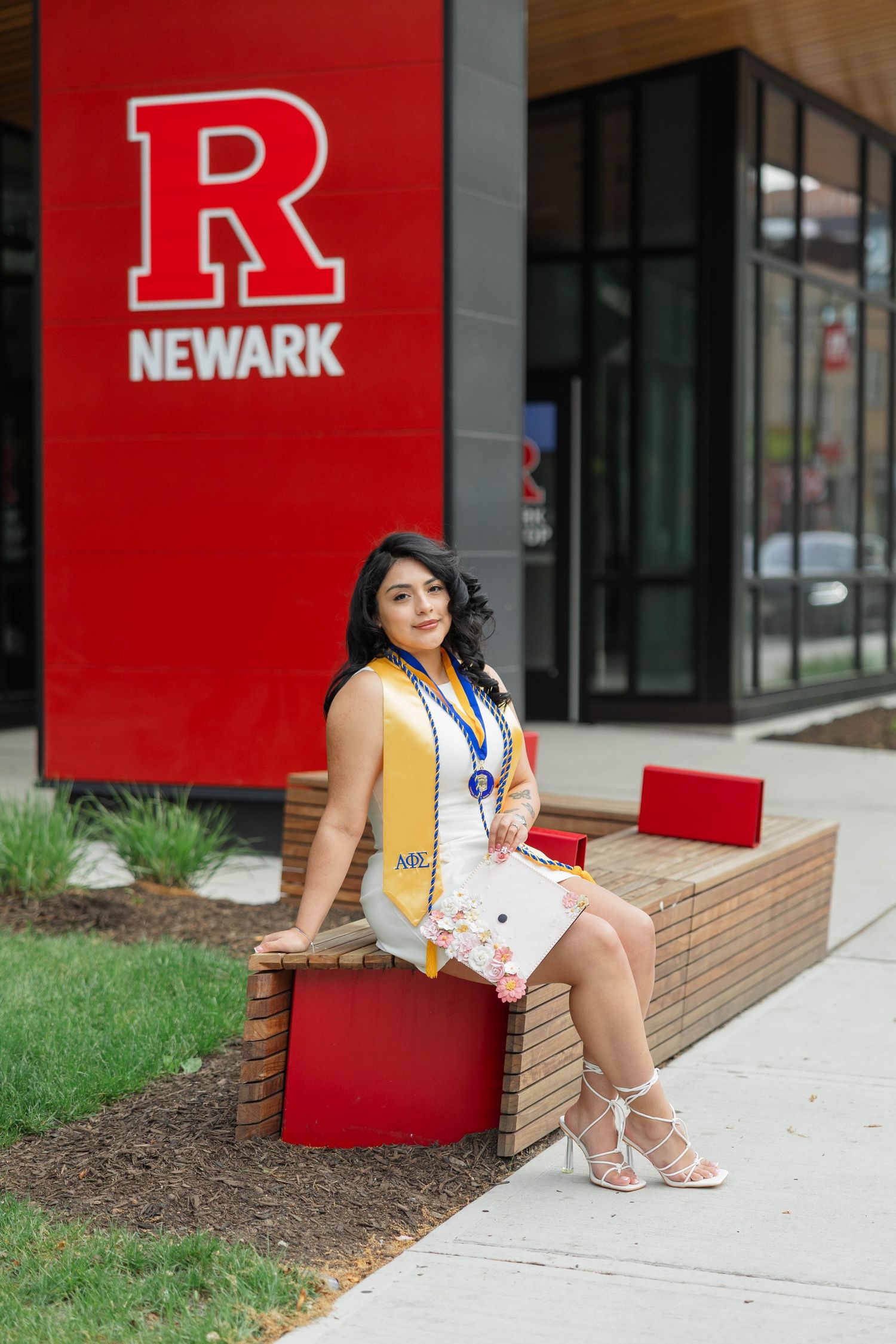 Rutgers Newark graduate poses in white dress with gold sash and medal on campus bench outside university building.