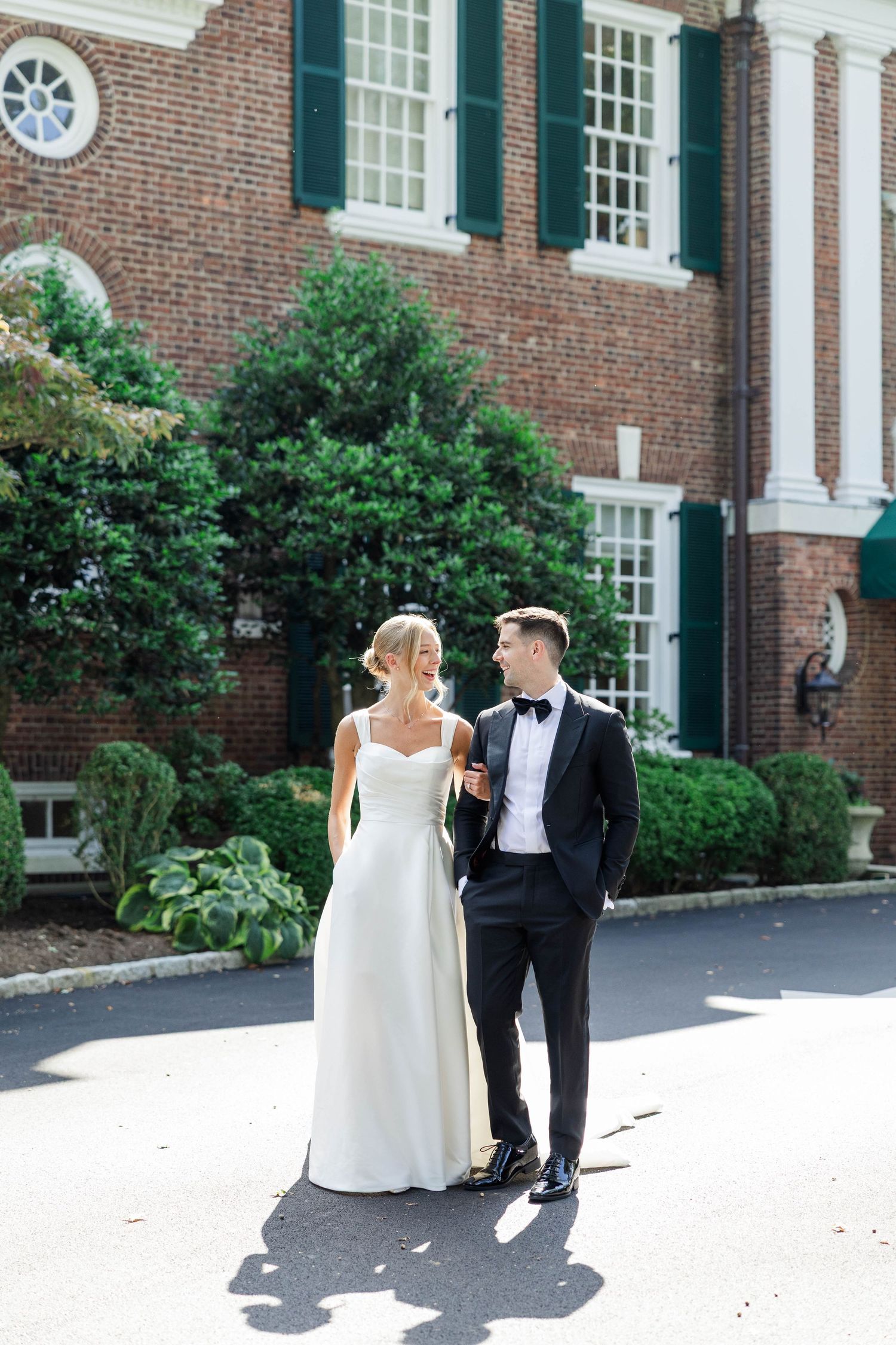 A couple walks together along a brick building with white columns and green shutters in formal wedding attire.
