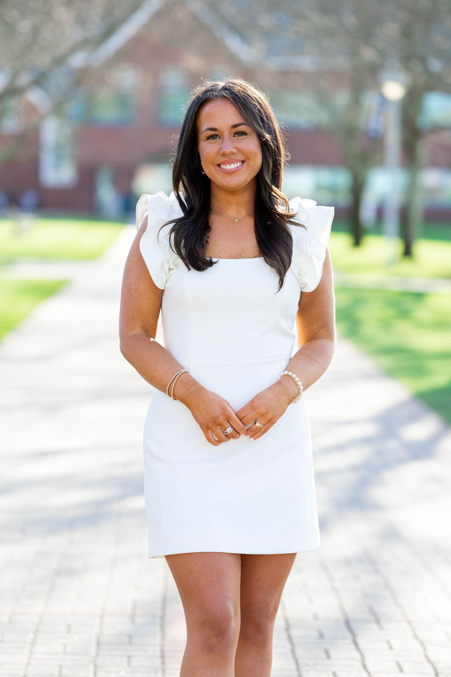 Smiling person in a white summer dress stands on a brick path with green grass and buildings in the background.