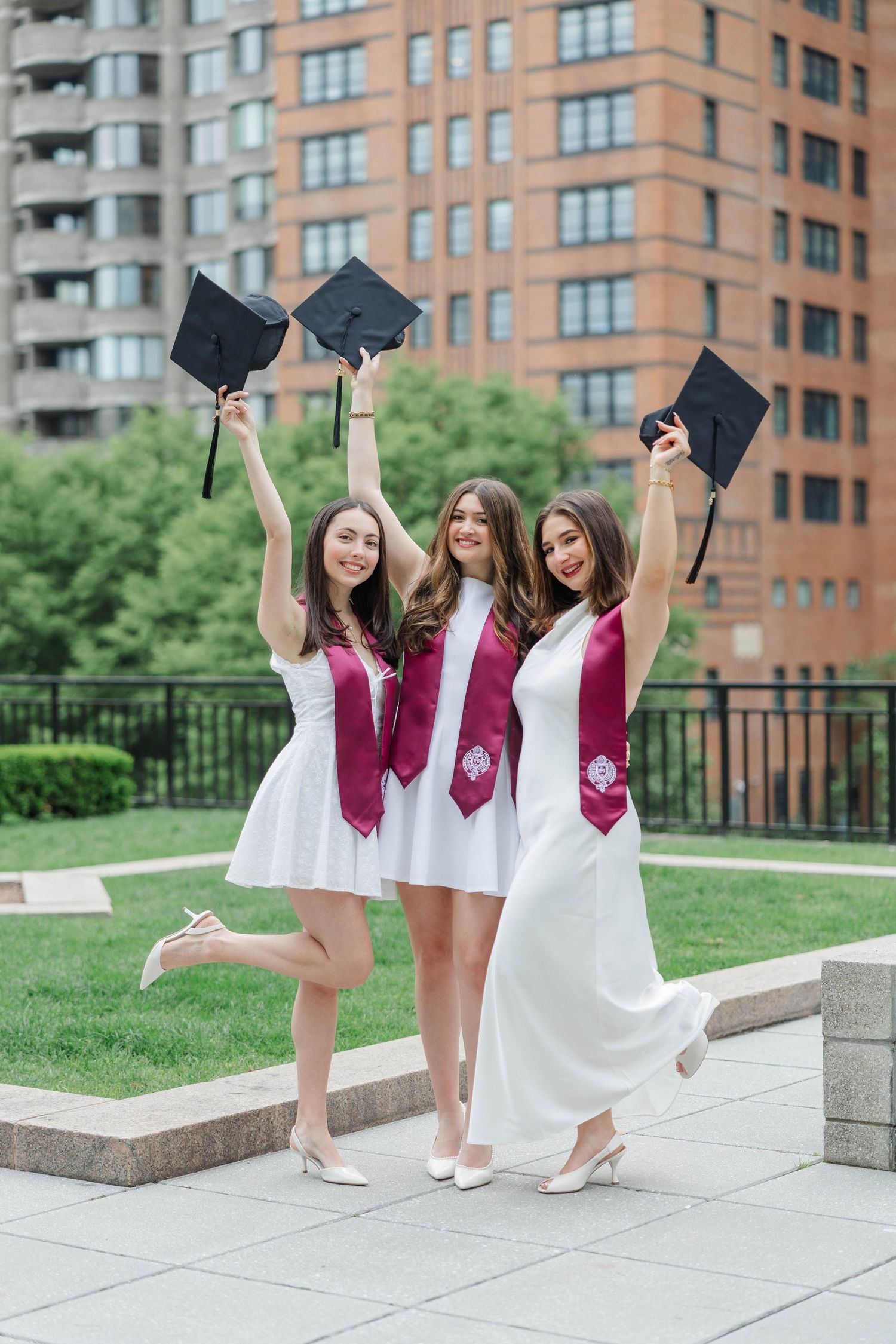 Three women in white dresses and maroon graduation stoles celebrate outdoors, raising caps in the air.