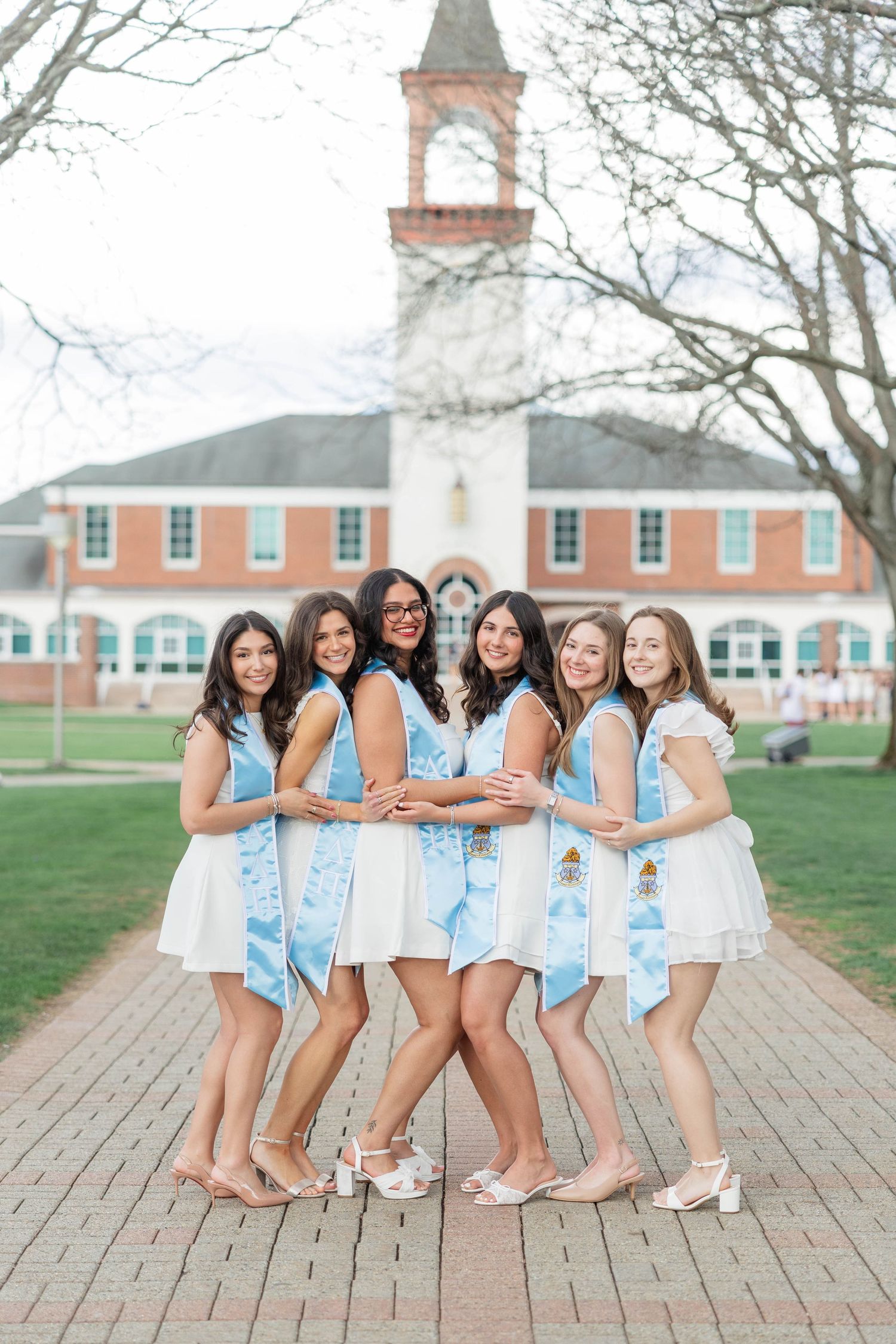 Six young women in white dresses and blue graduation stoles pose together on a brick path in front of a brick building.