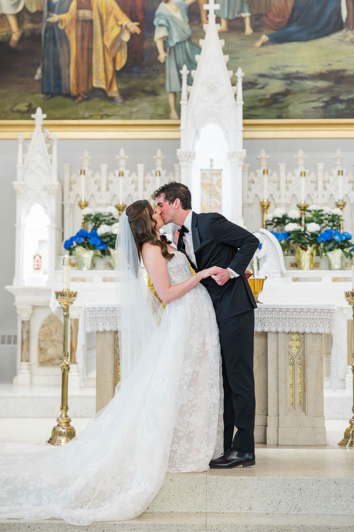 Bride and groom share first kiss at ornate white church altar, bride in lace gown with veil, groom in black tuxedo.