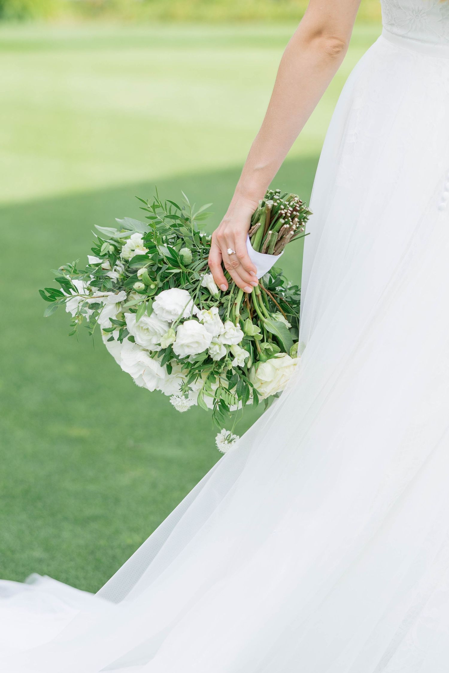 Bride in white gown holding lush bouquet of white flowers and greenery on a sunny golf course.