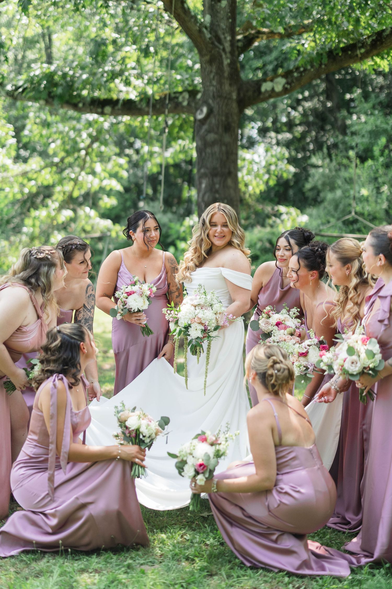 Bride in white gown surrounded by bridesmaids in dusty rose dresses holding floral bouquets outdoors under a tree.