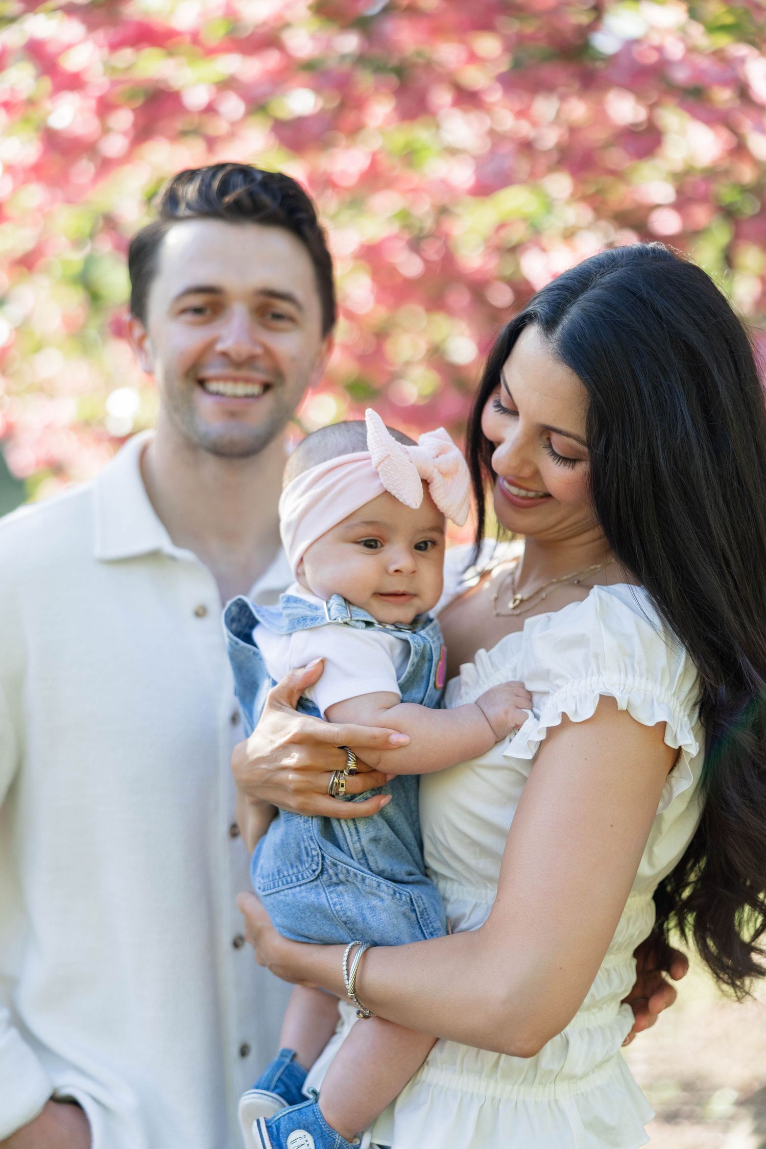 Happy young family with smiling parents holding their baby girl in a pink bow outdoors near blooming pink trees.