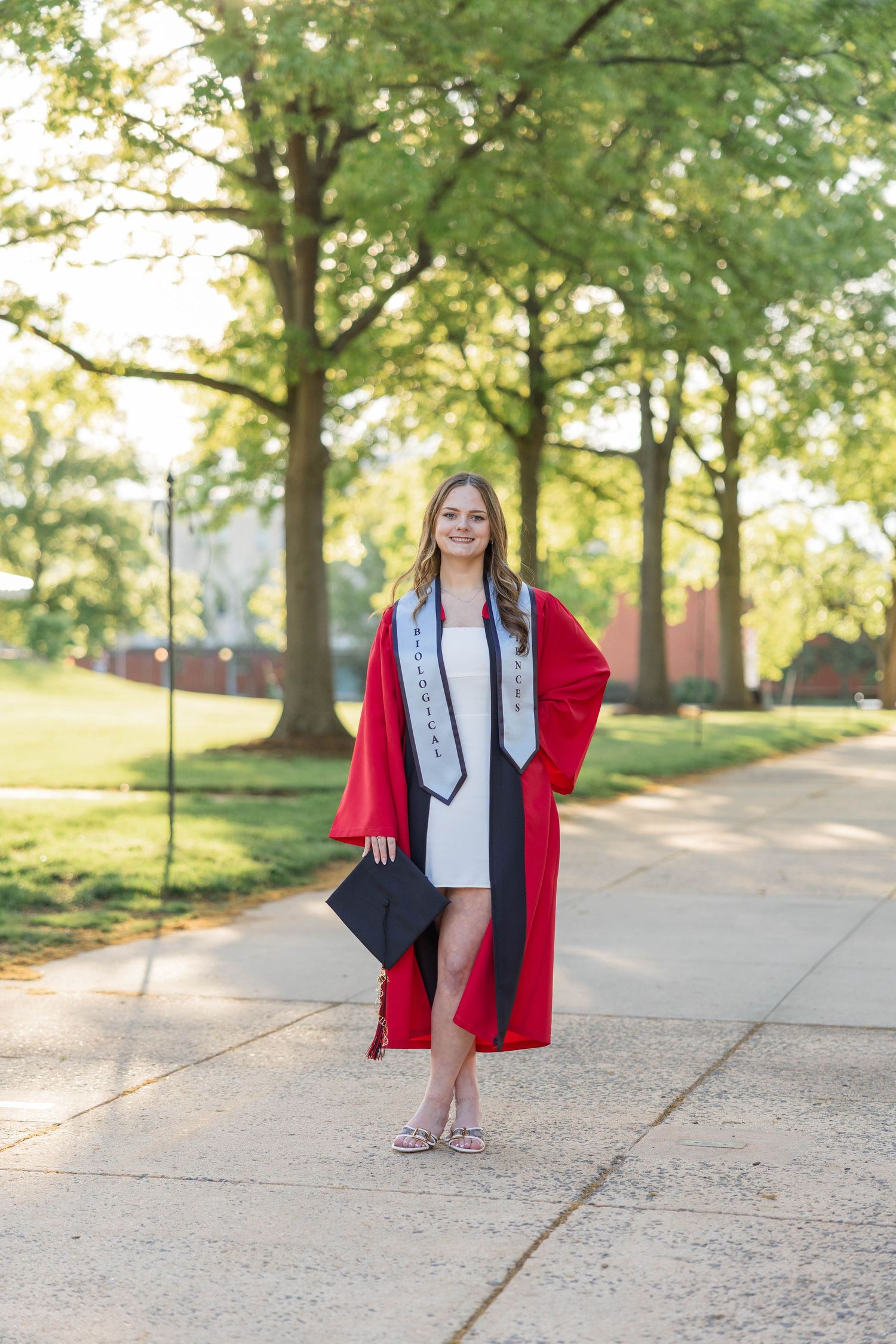 Young woman in red graduation gown and white dress holding cap on tree-lined campus walkway.