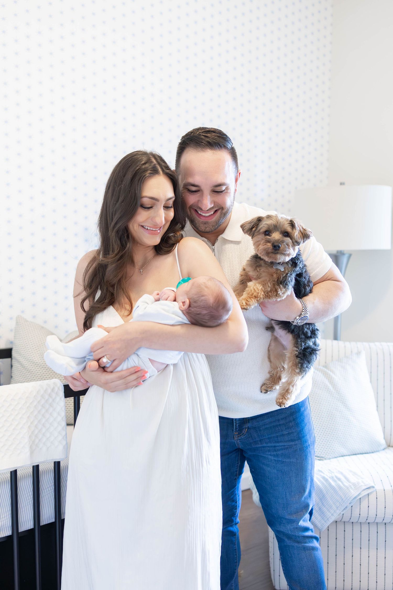 A couple in white and denim holds their small dog while posing together in a bright indoor setting.