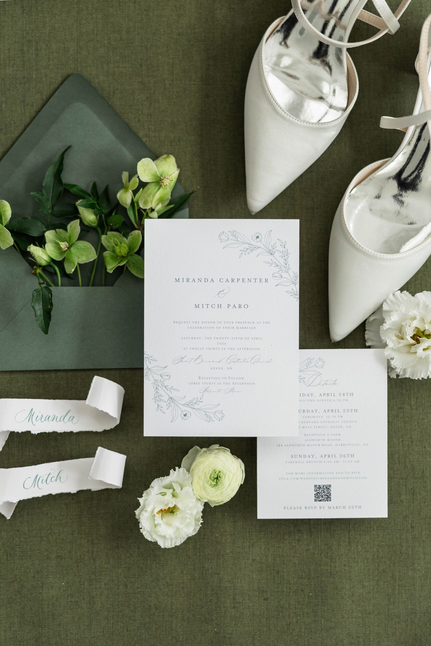 Elegant wedding invitation suite with floral details, white heels, green envelope, and ribbon name tags on sage linen.