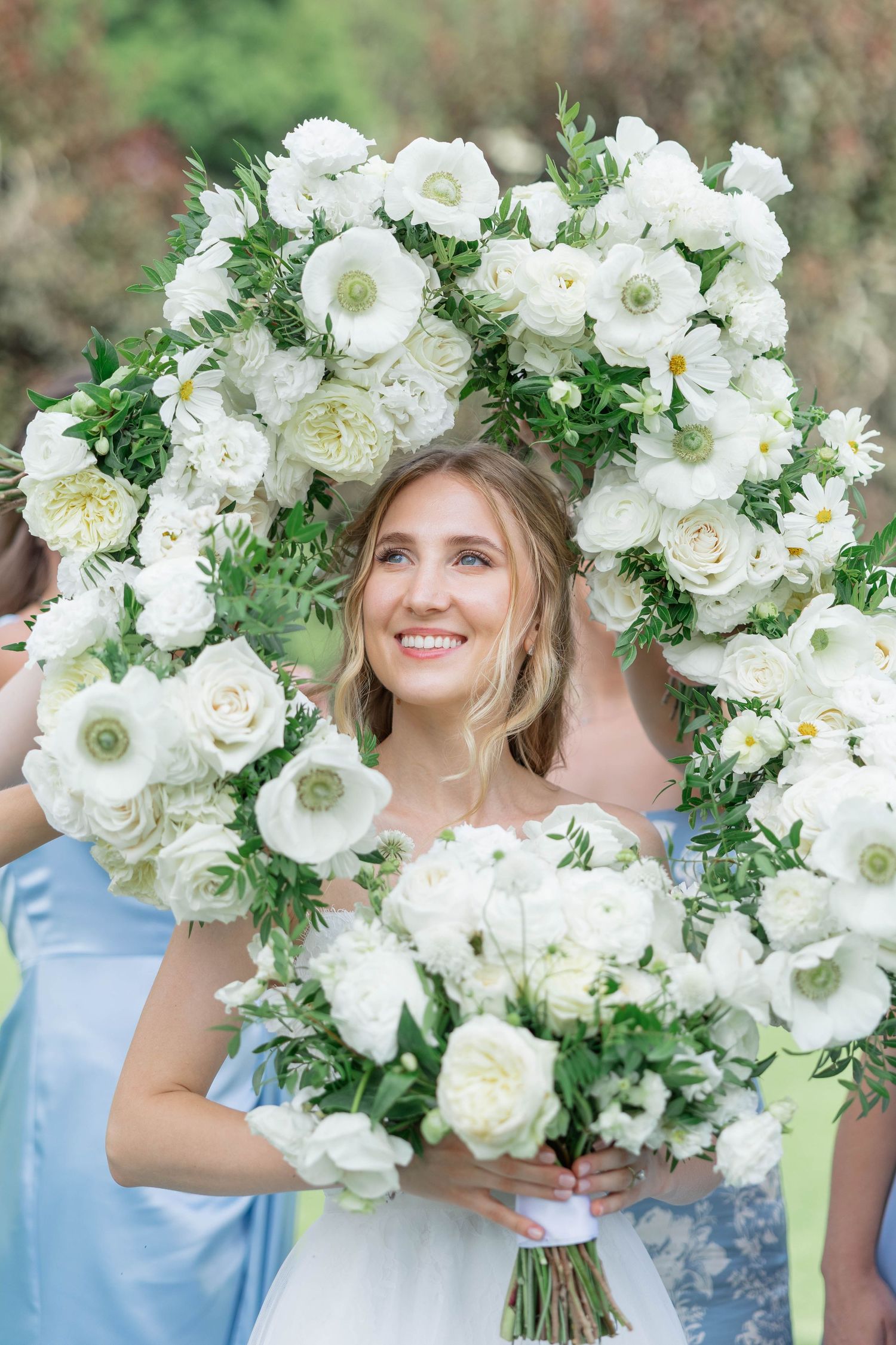 Smiling bride framed by white floral bouquet arch holding white roses and ranunculus at outdoor wedding.