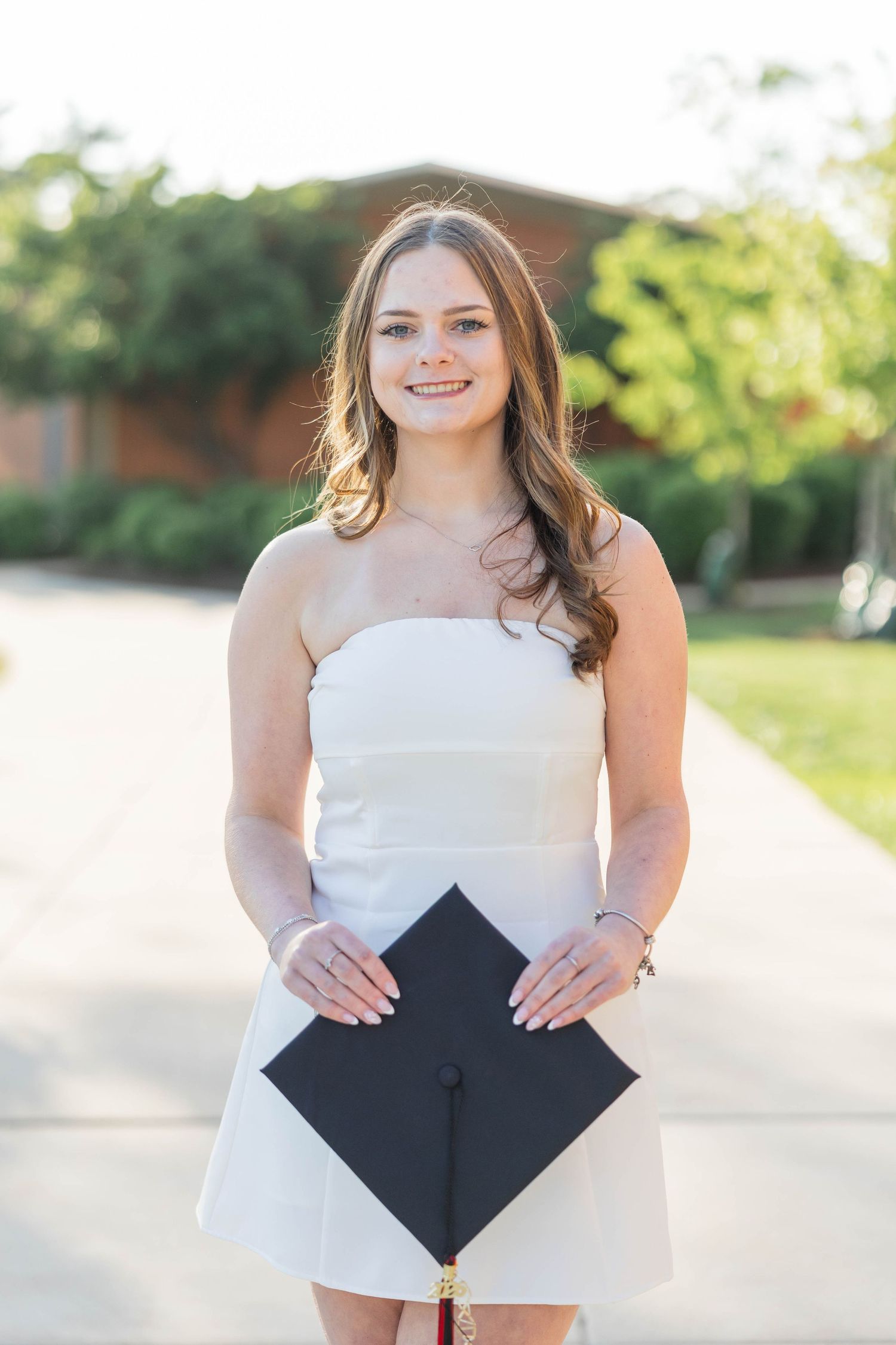 Young woman in white strapless dress smiling outdoors, holding black graduation cap during commencement photo session.