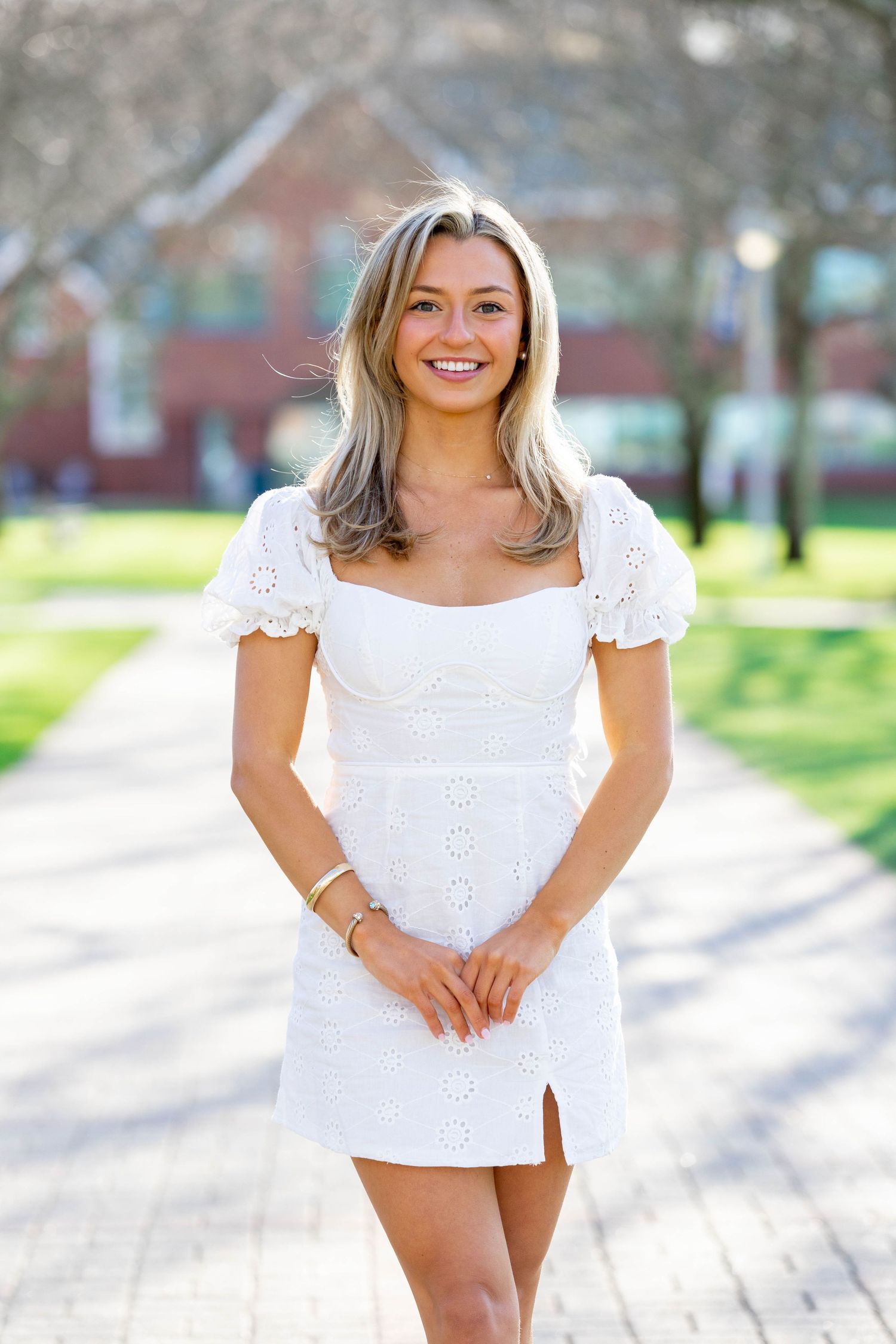 Young woman in white eyelet mini dress smiling outdoors on a sunny campus pathway with green grass.