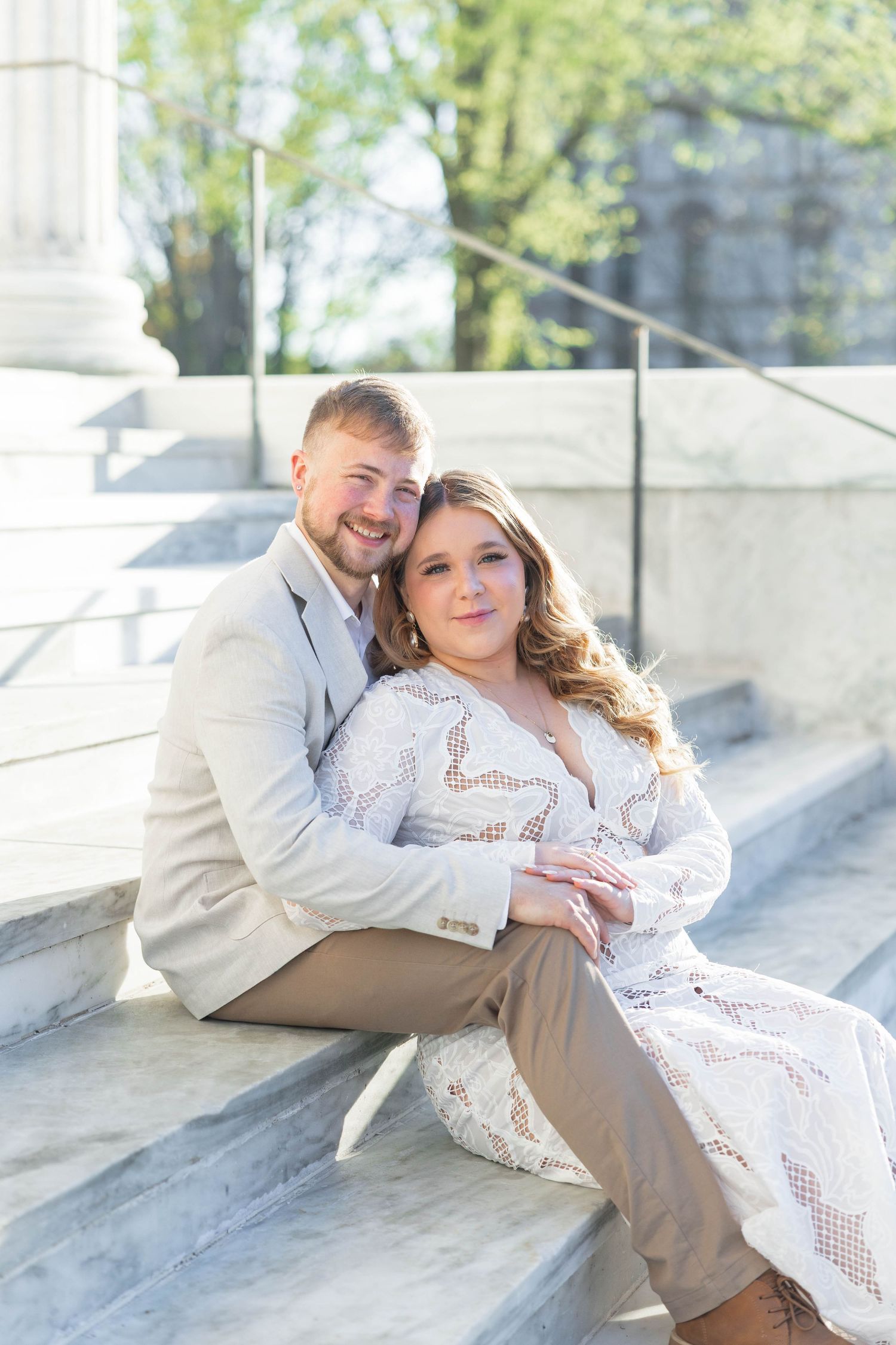 A couple sits on marble steps, the man in a tan suit embracing a woman in a white lace dress, smiling warmly.