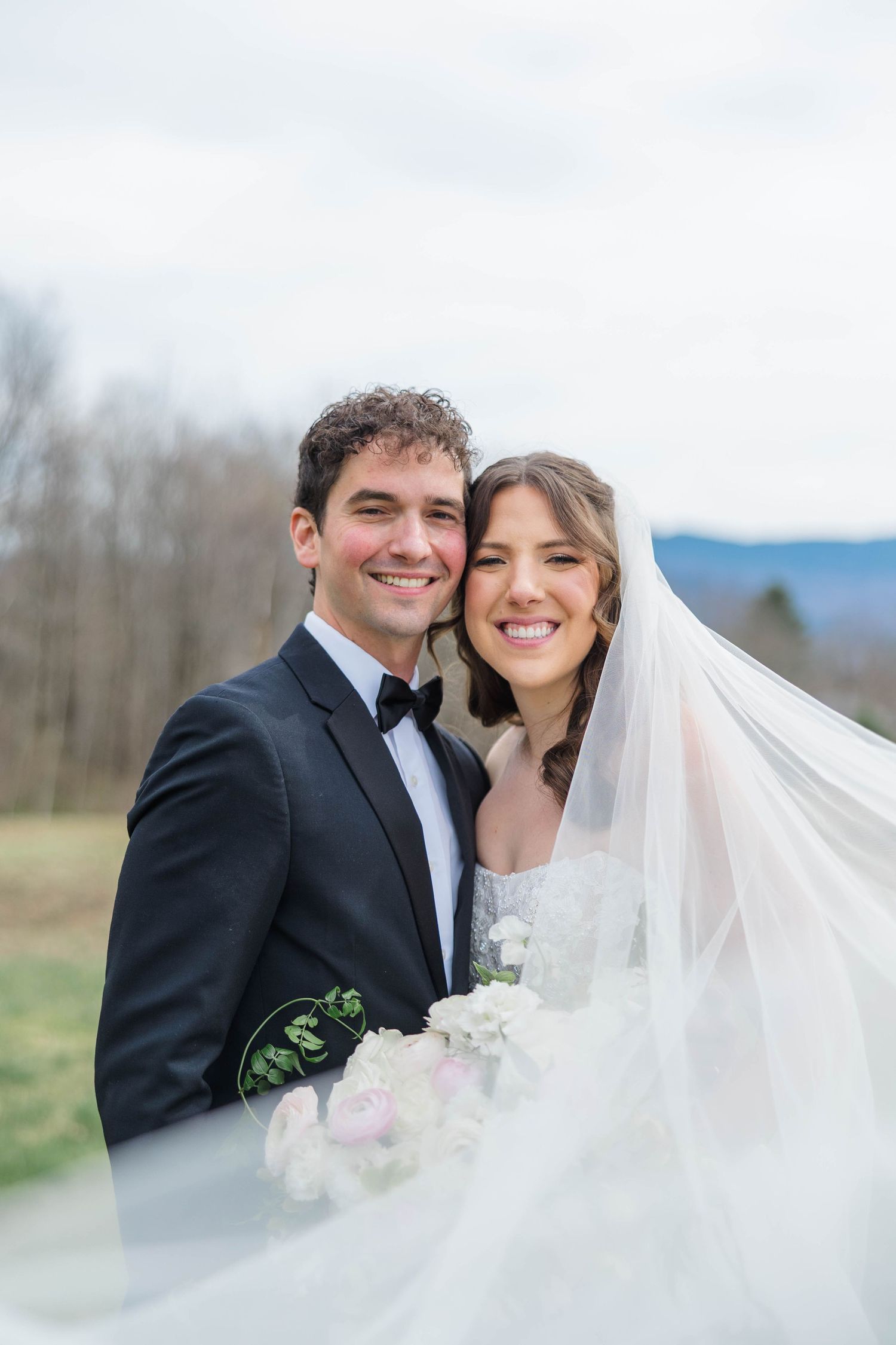 Smiling bride and groom pose outdoors, bride wearing veil and holding bouquet, groom in black tuxedo.