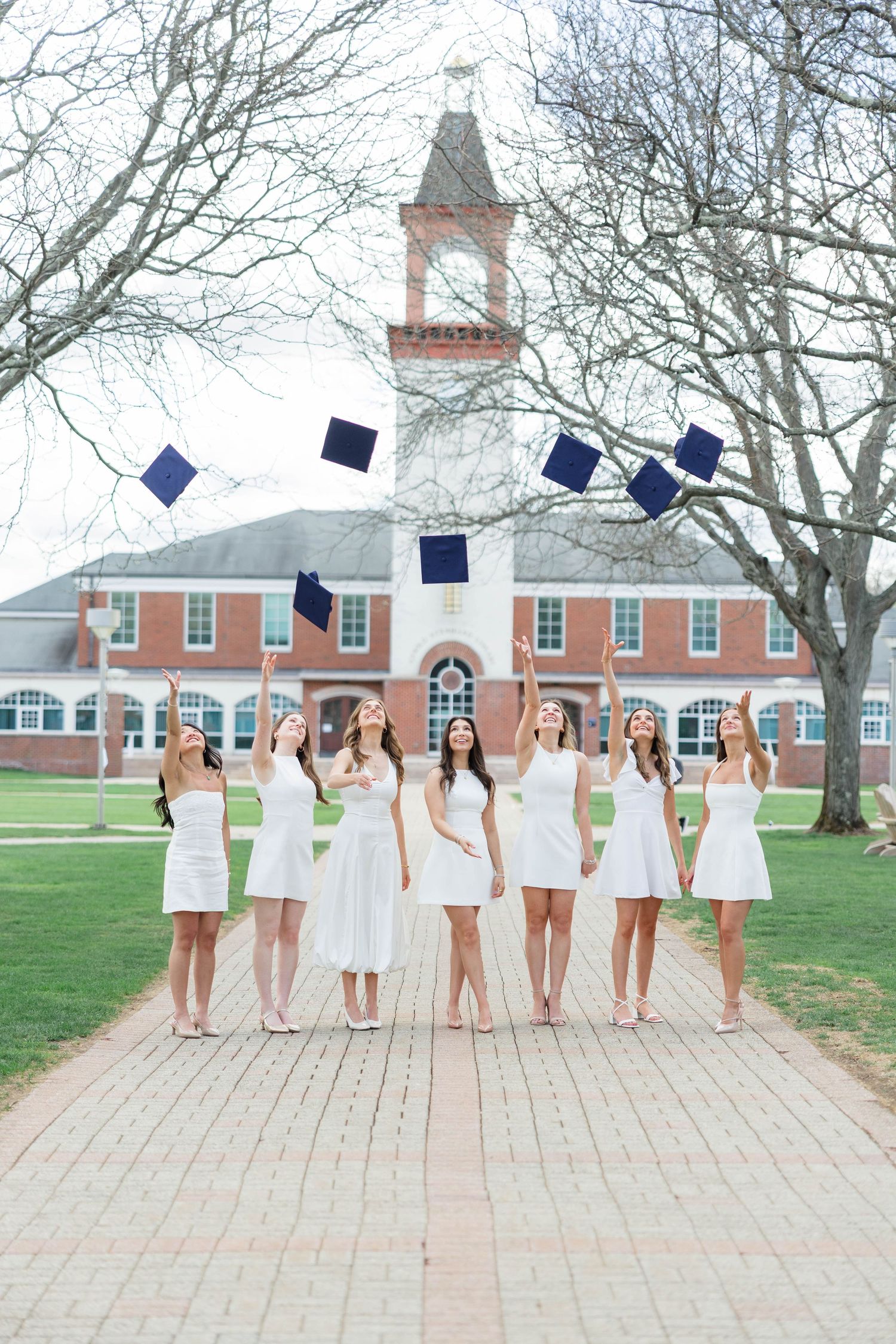 Seven women in white dresses toss navy graduation caps in the air on a brick path outside a red brick university building.