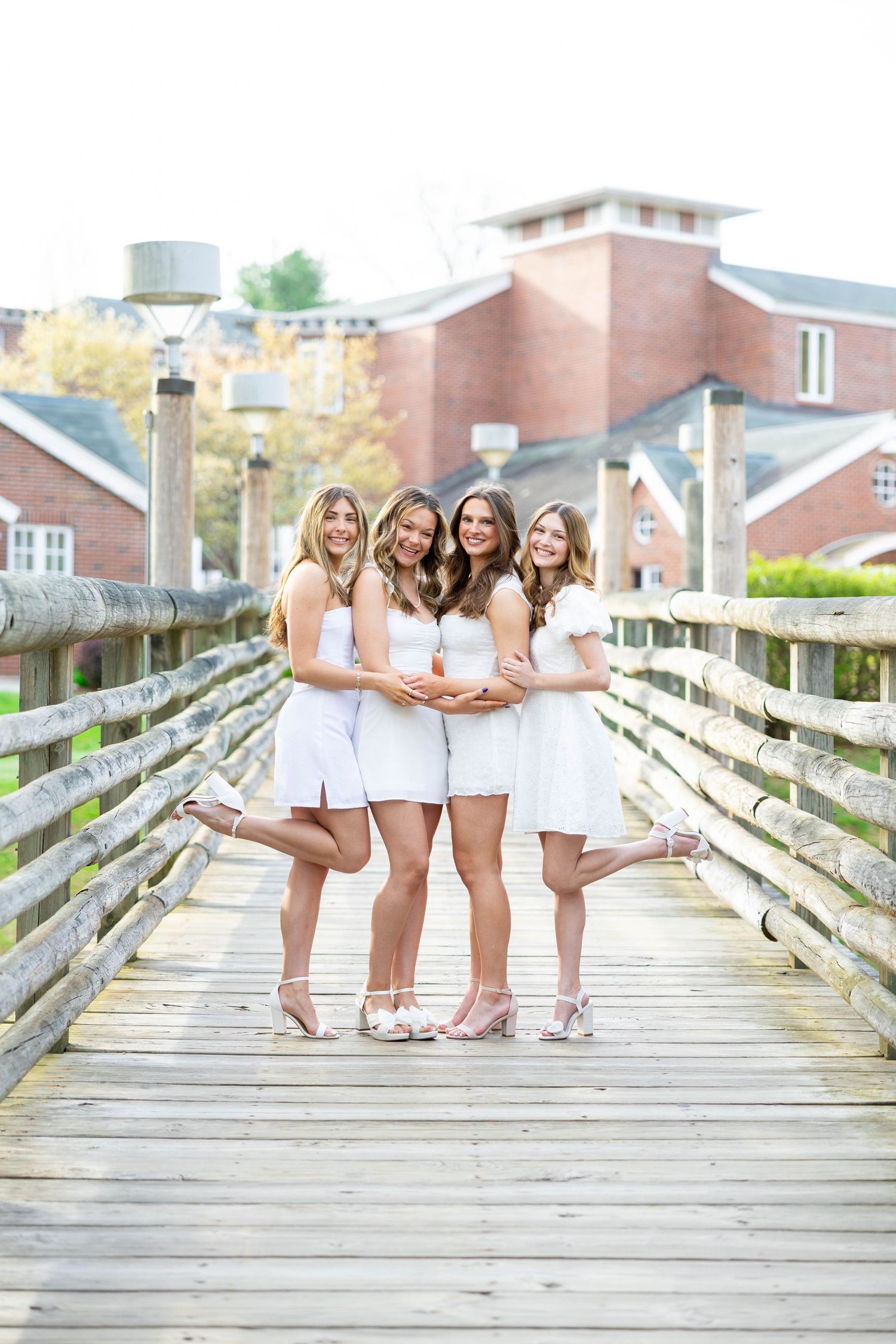 Four graduates in white dresses pose playfully on wooden bridge during photo shoot.