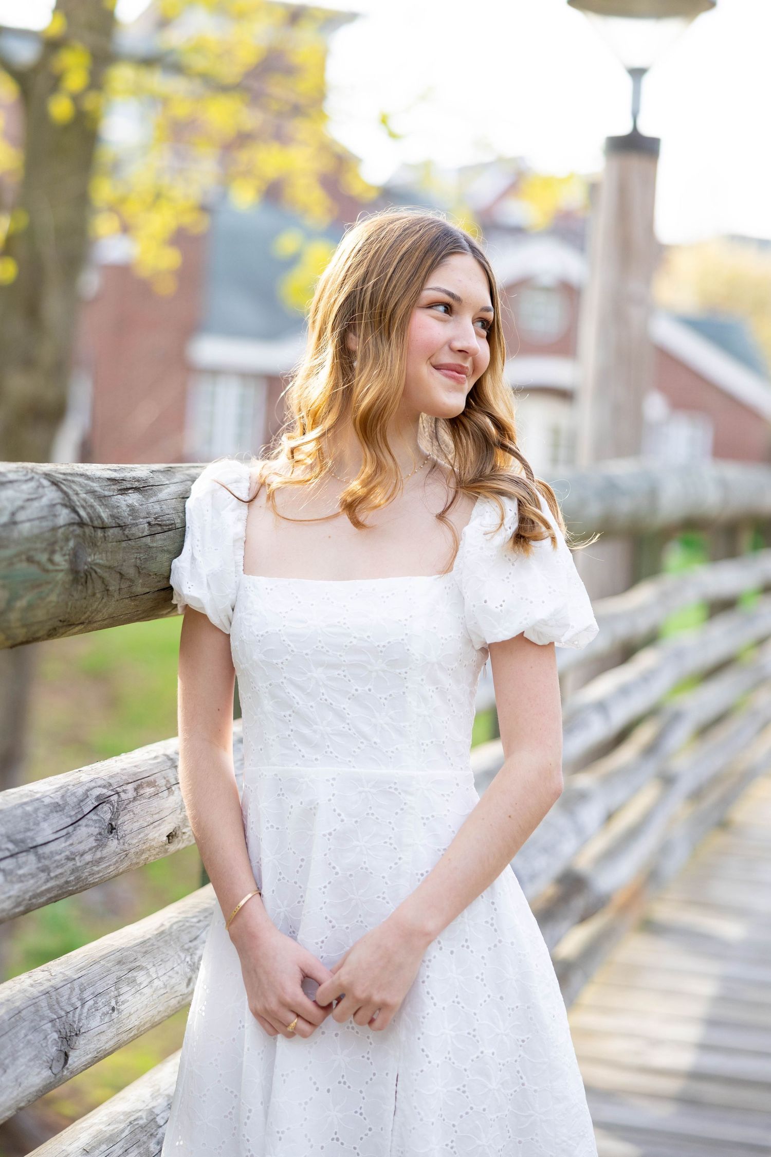 Woman in white eyelet dress poses by rustic wooden fence during spring photo session.