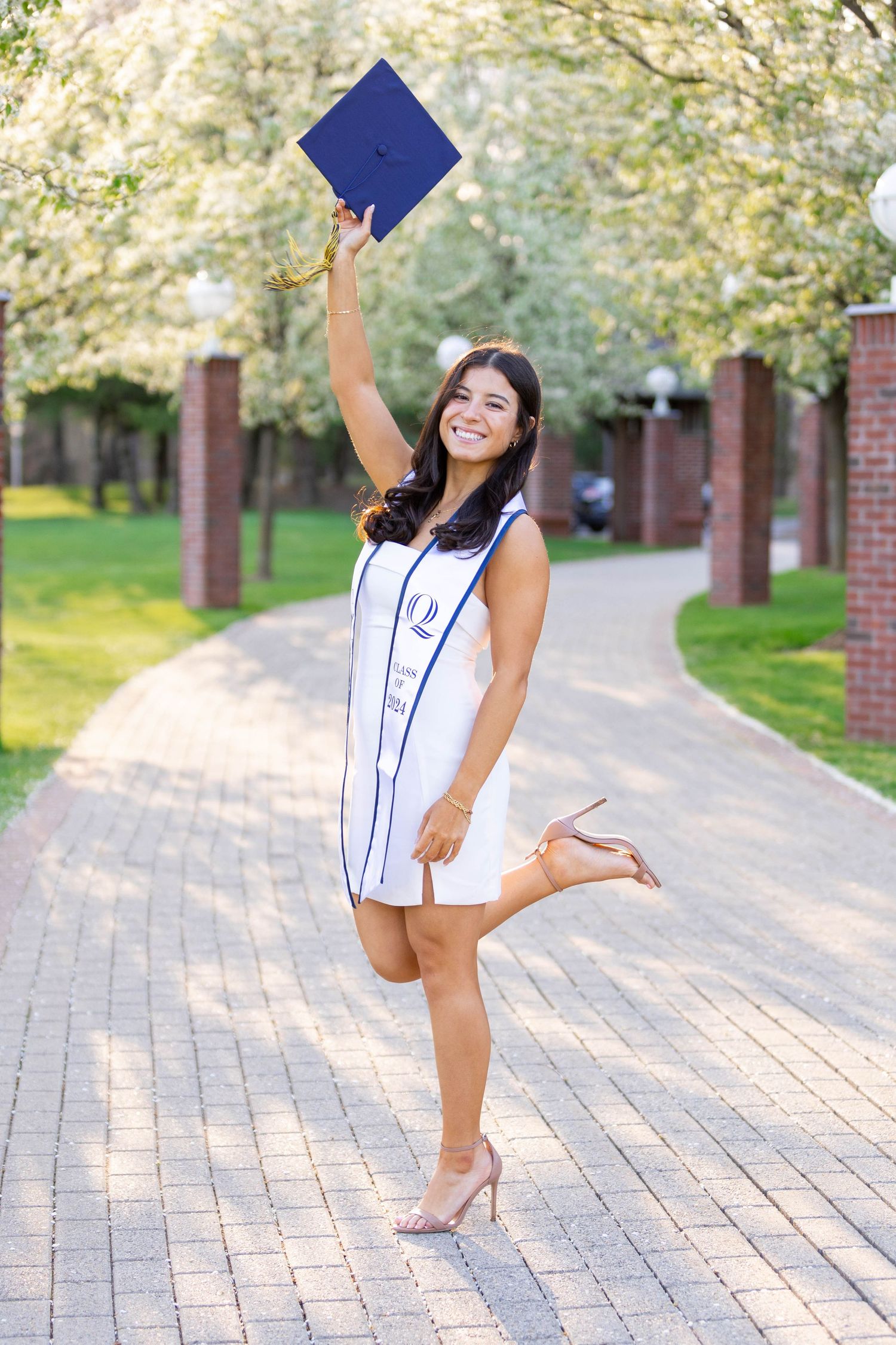 Graduate in white dress and blue sash celebrating with diploma on campus pathway.