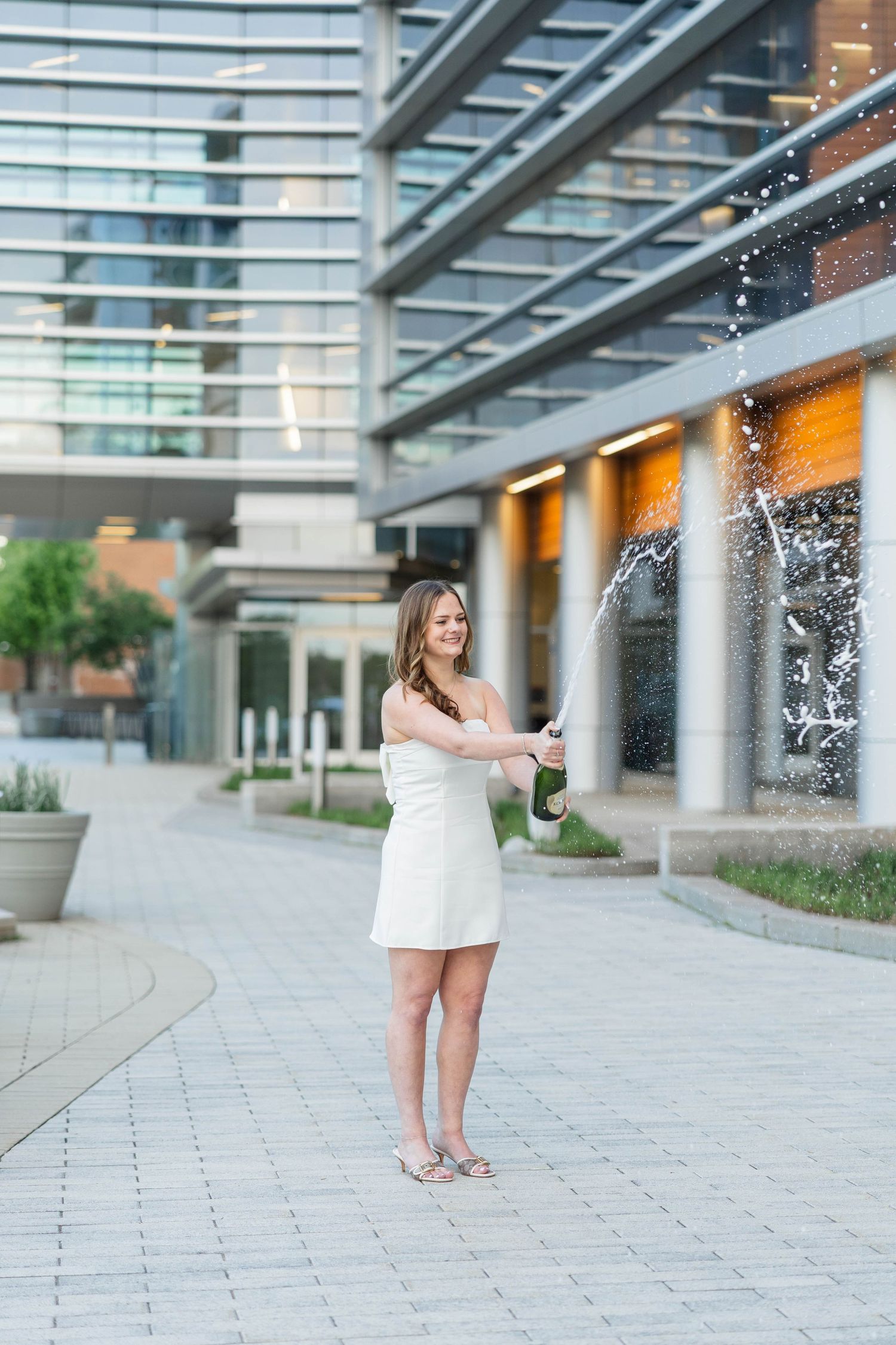Woman in white dress popping champagne bottle outdoors near modern glass building on paved walkway.