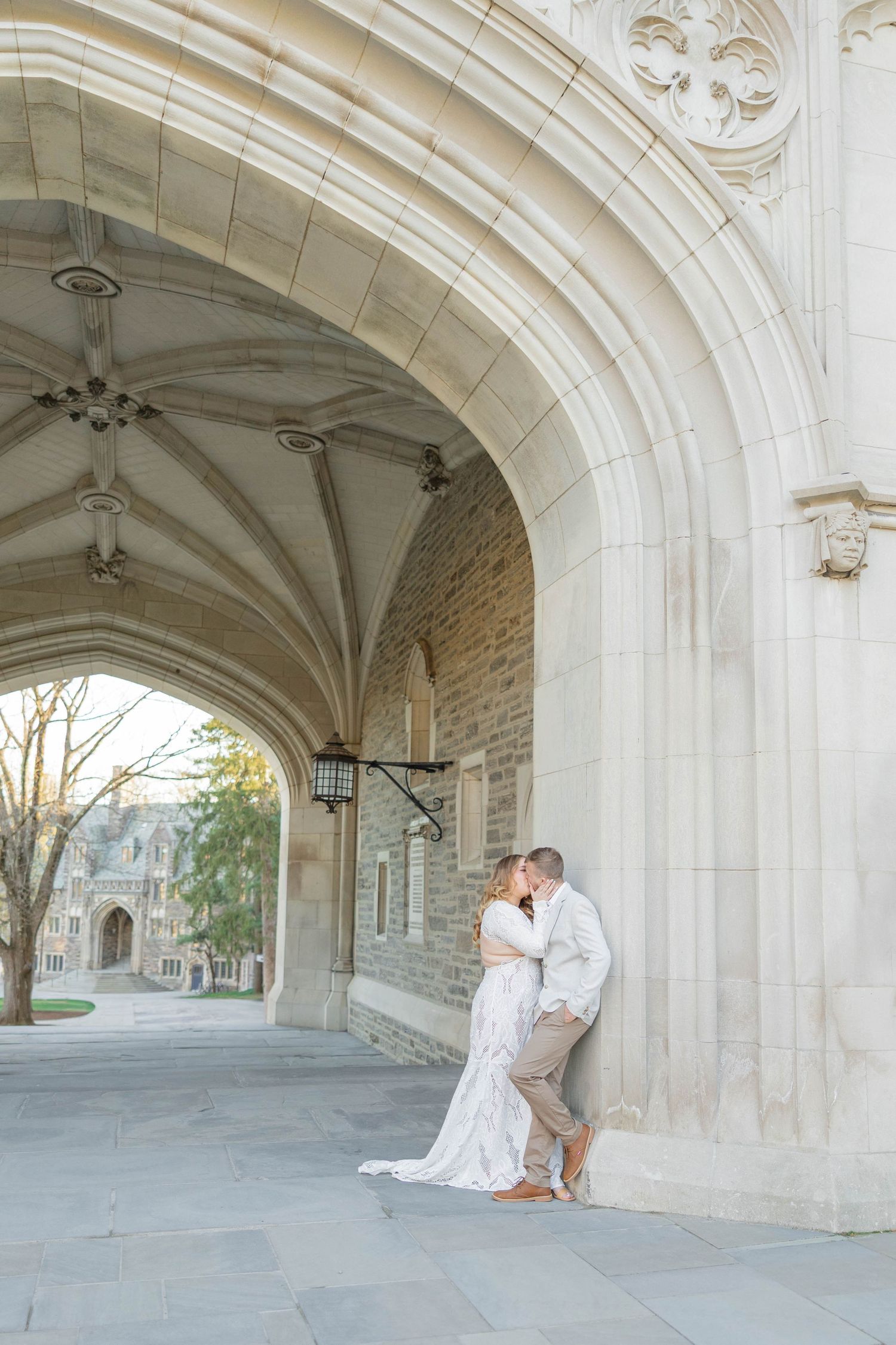 Bride and groom share a romantic kiss under a Gothic stone archway, bride in lace gown, groom in tan suit.