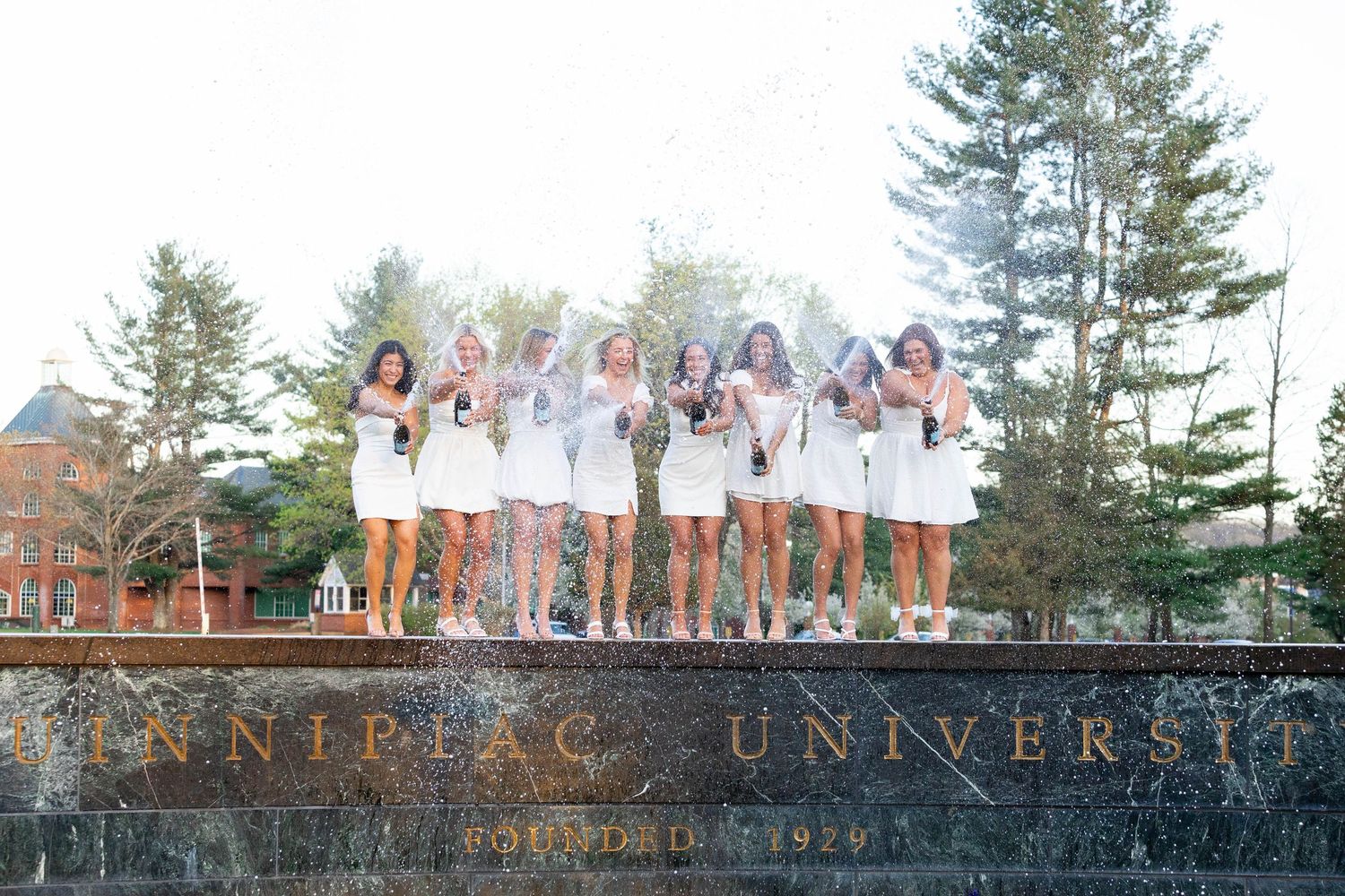 Group of women in white dresses celebrating on Quinnipiac University sign, founded 1929.