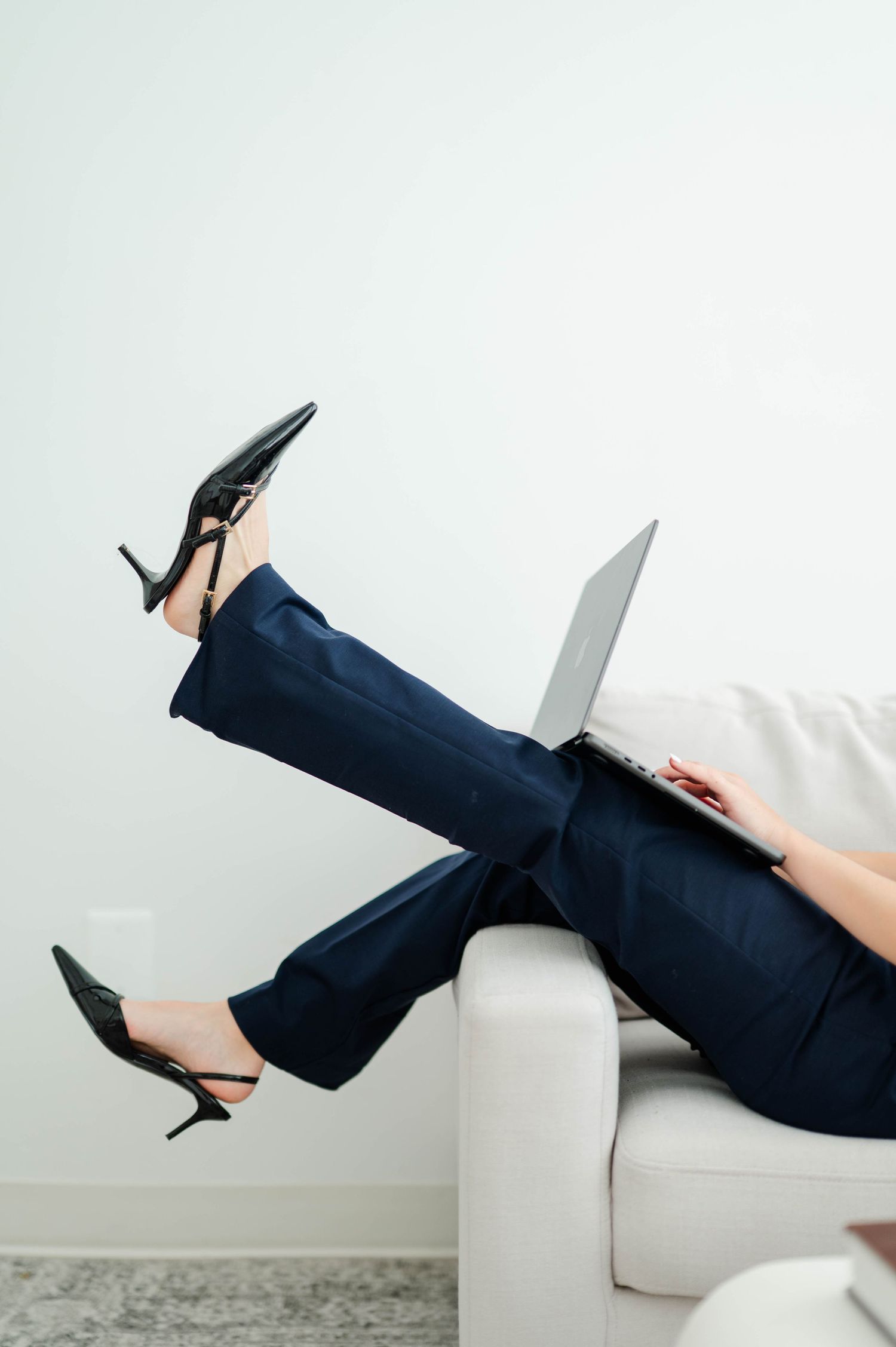 Professional woman in navy pants and heels relaxes on white sofa while working on laptop computer.