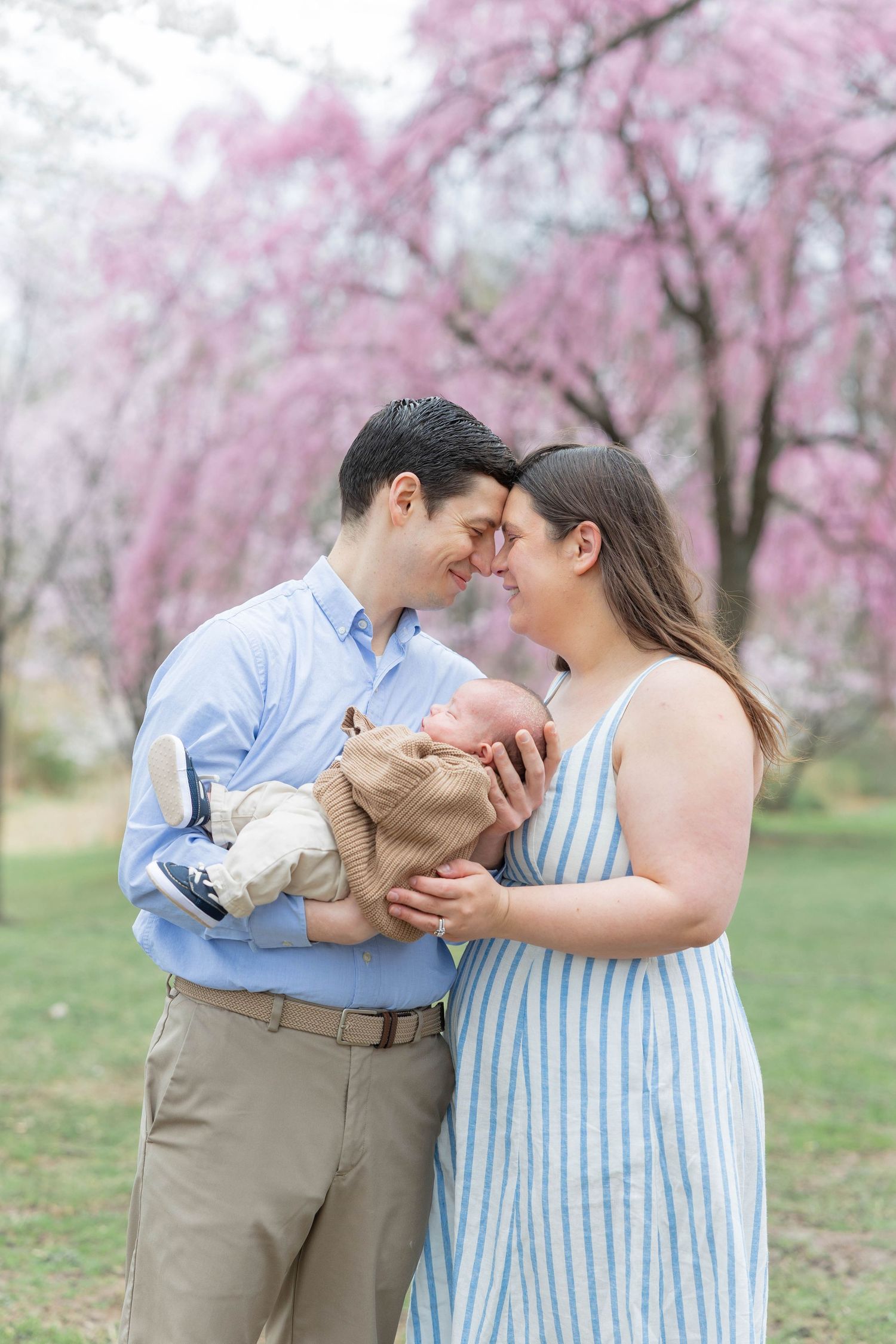 A couple shares a tender moment with their newborn baby outdoors amid blooming pink cherry blossom trees in spring.