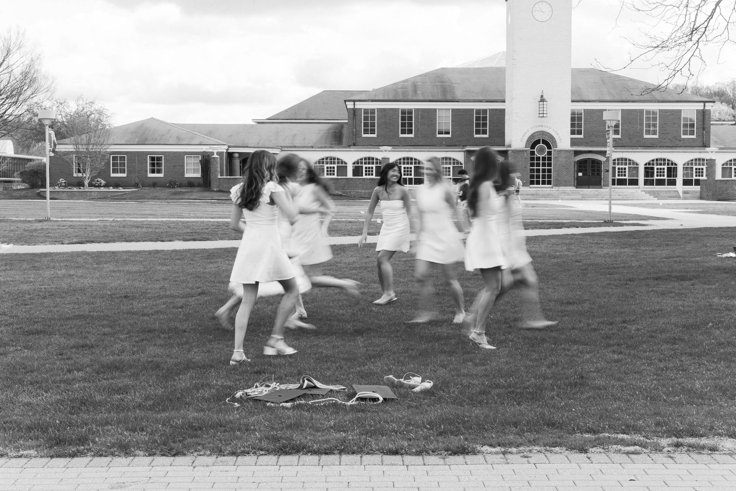 Black and white photo of girls in white dresses playing on a grass lawn near a brick building.