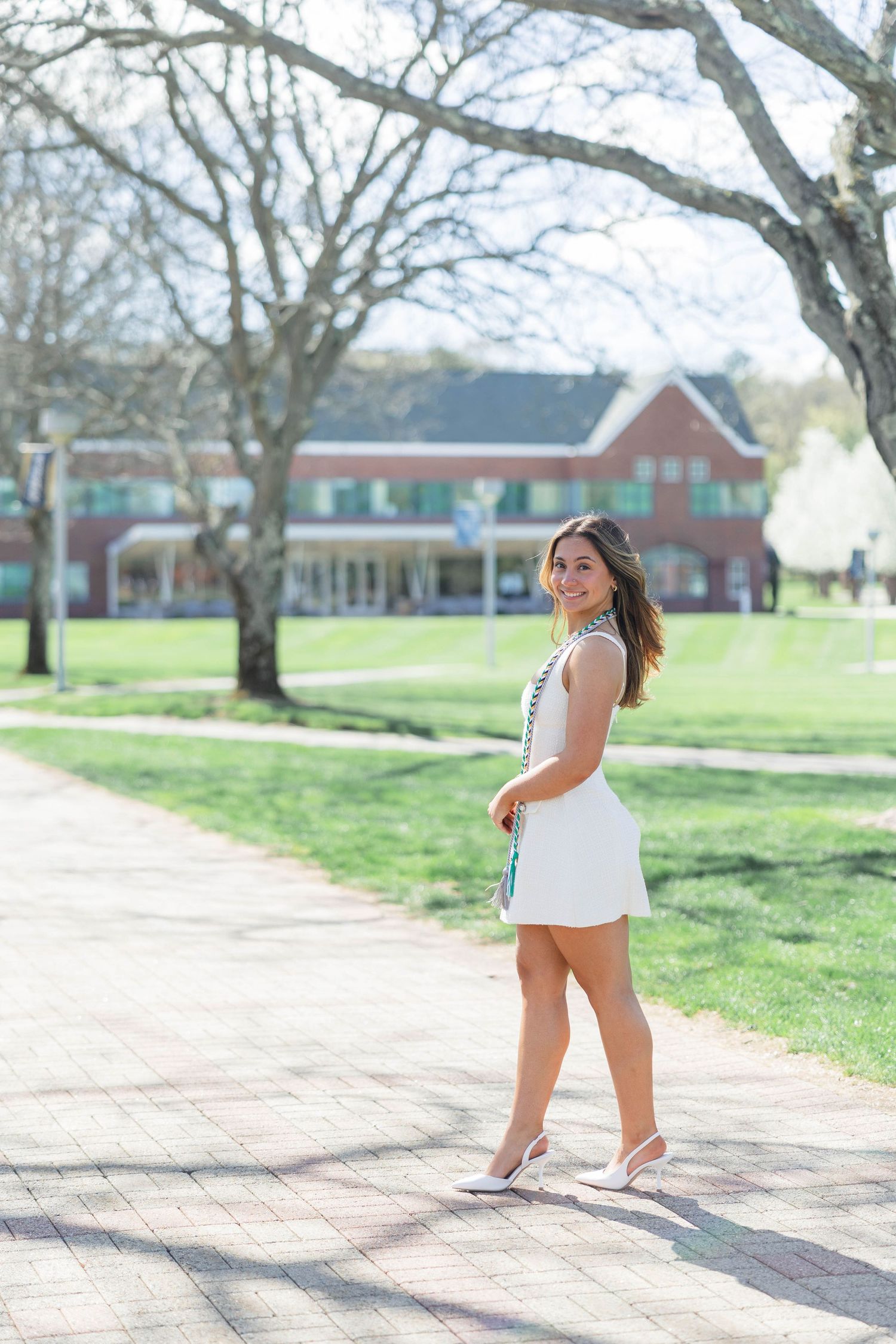 Young woman in white mini dress and heels walking on brick path in sunny campus setting with brick building behind her.