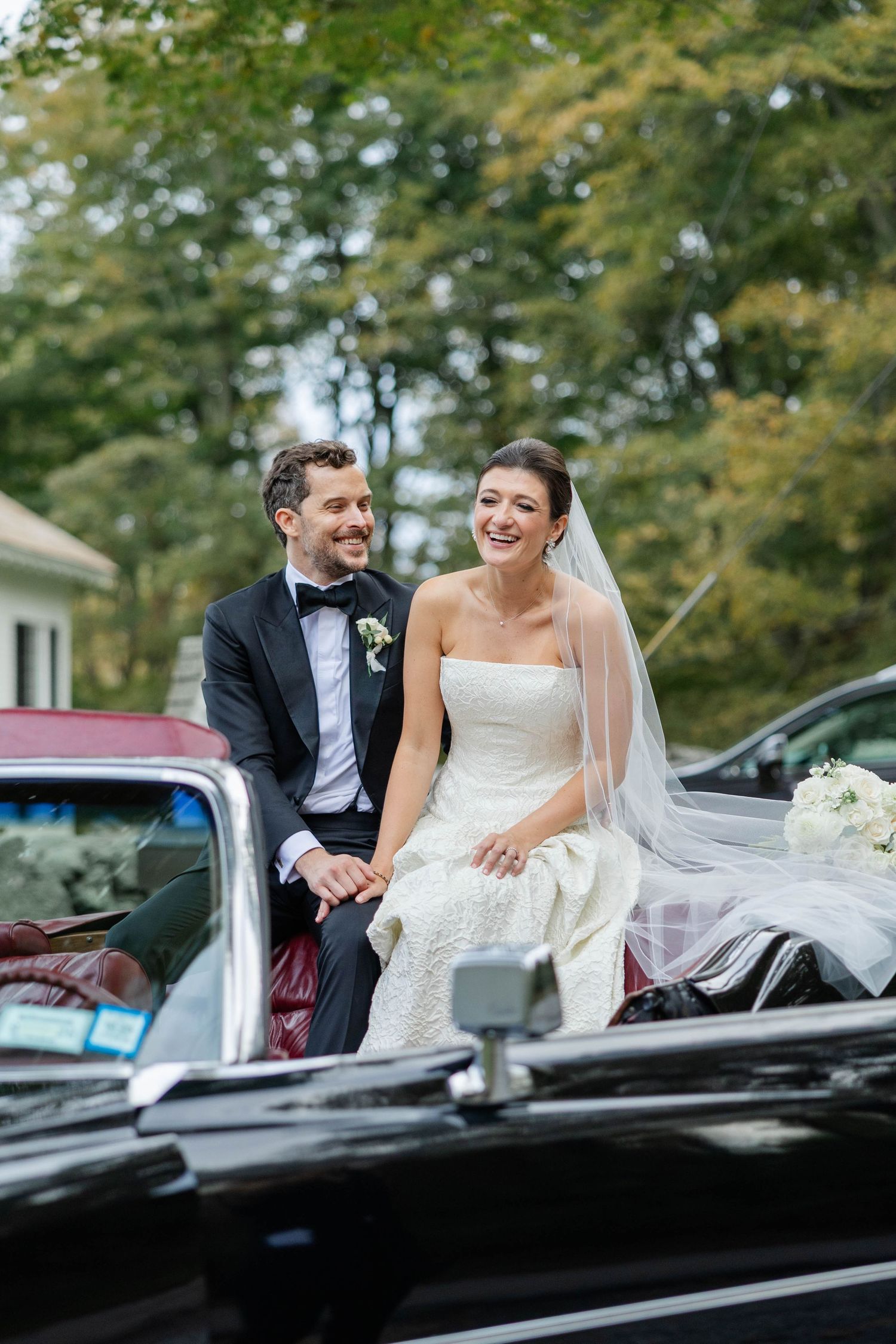Happy couple sits on vintage black car after wedding ceremony, surrounded by trees and natural light.