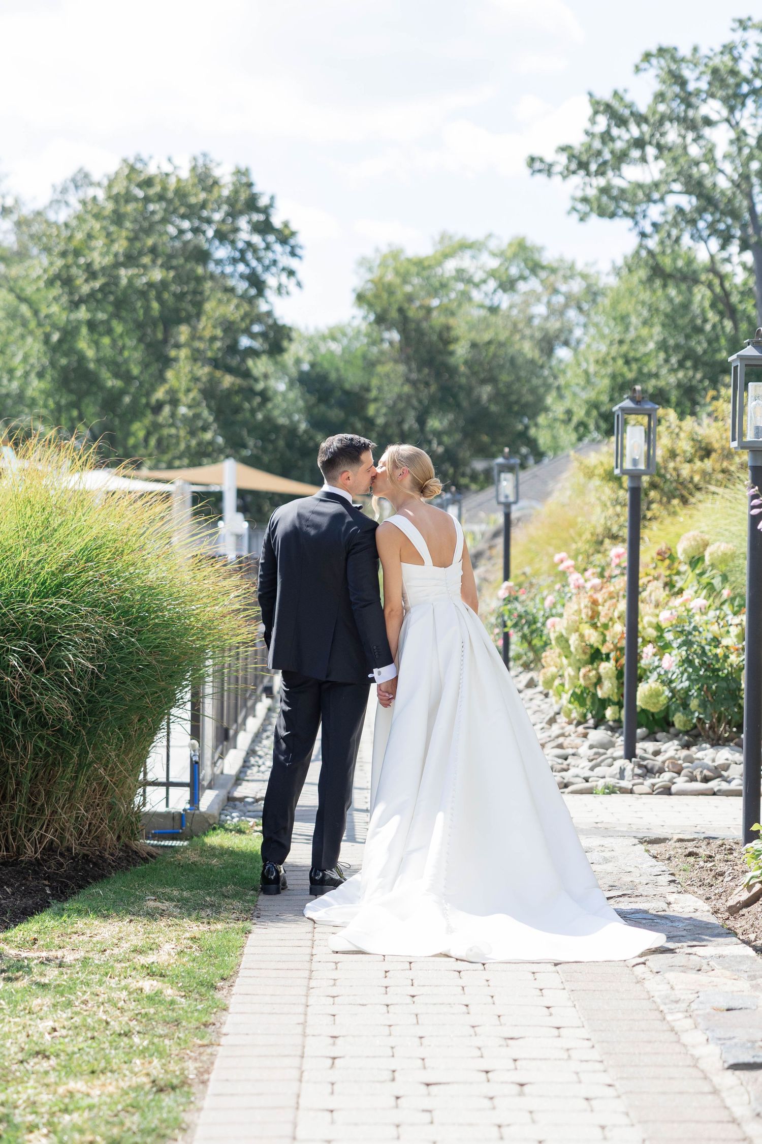Bride and groom sharing a kiss on a garden pathway, with lush greenery and flowers in the background on their wedding day.